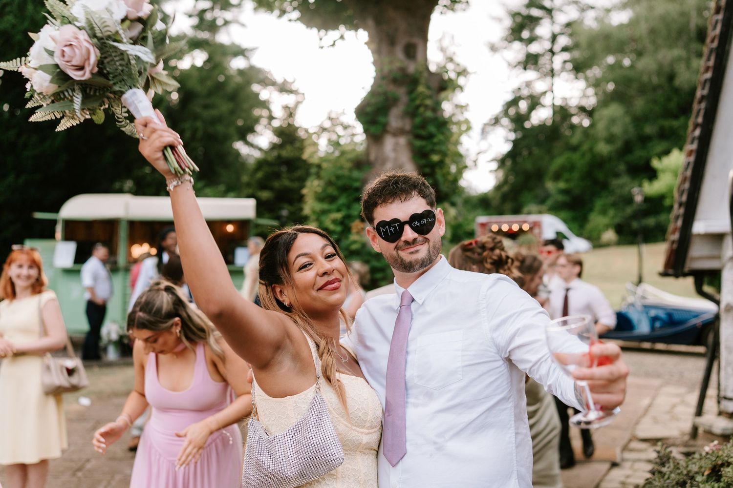 Excited wedding guest raises bouquet while posing with man in heart sunglasses at outdoor reception.