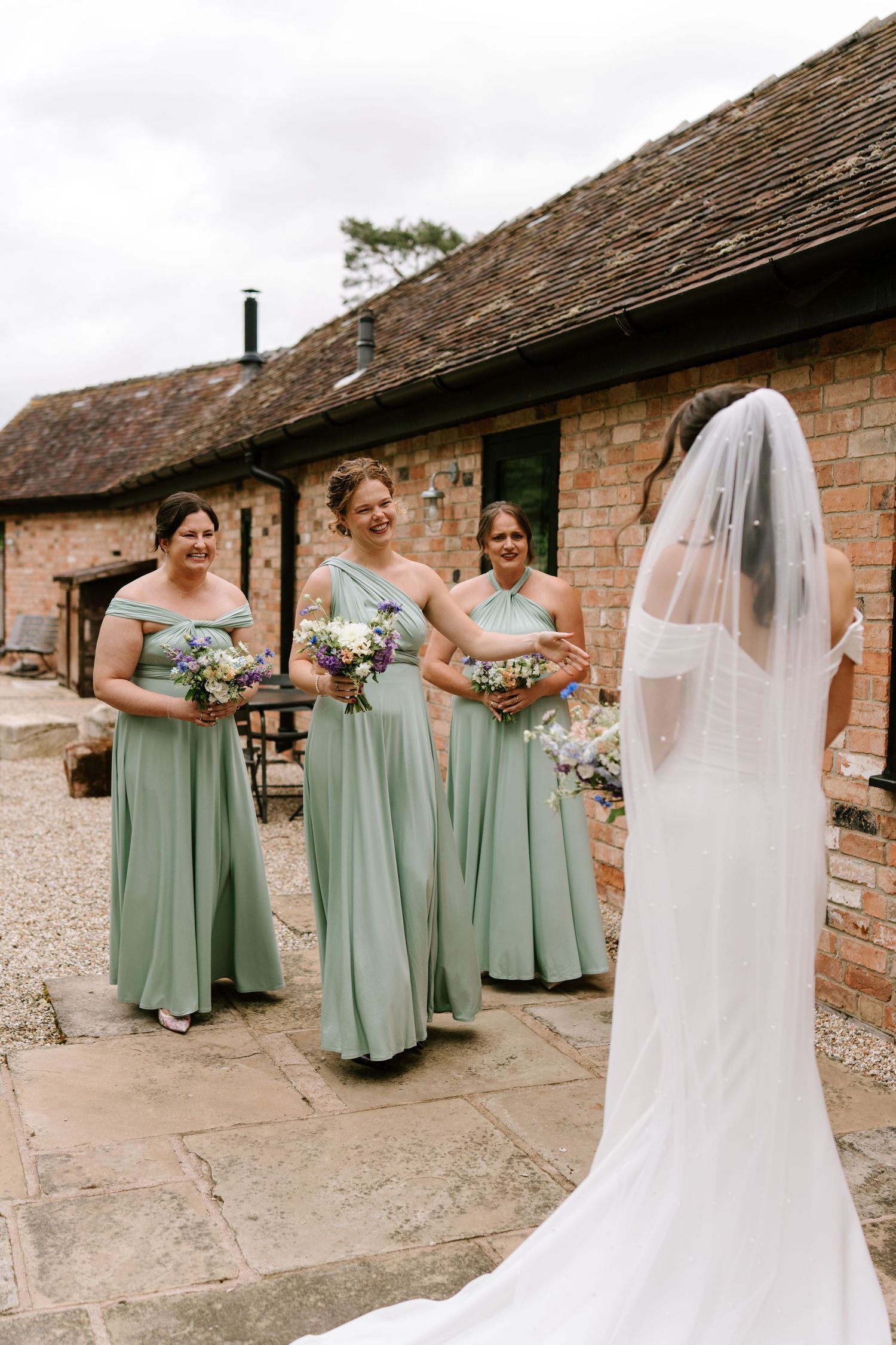 Three bridesmaids in sage green gowns holding bouquets smile at a bride in a white veil at a rustic barn wedding.