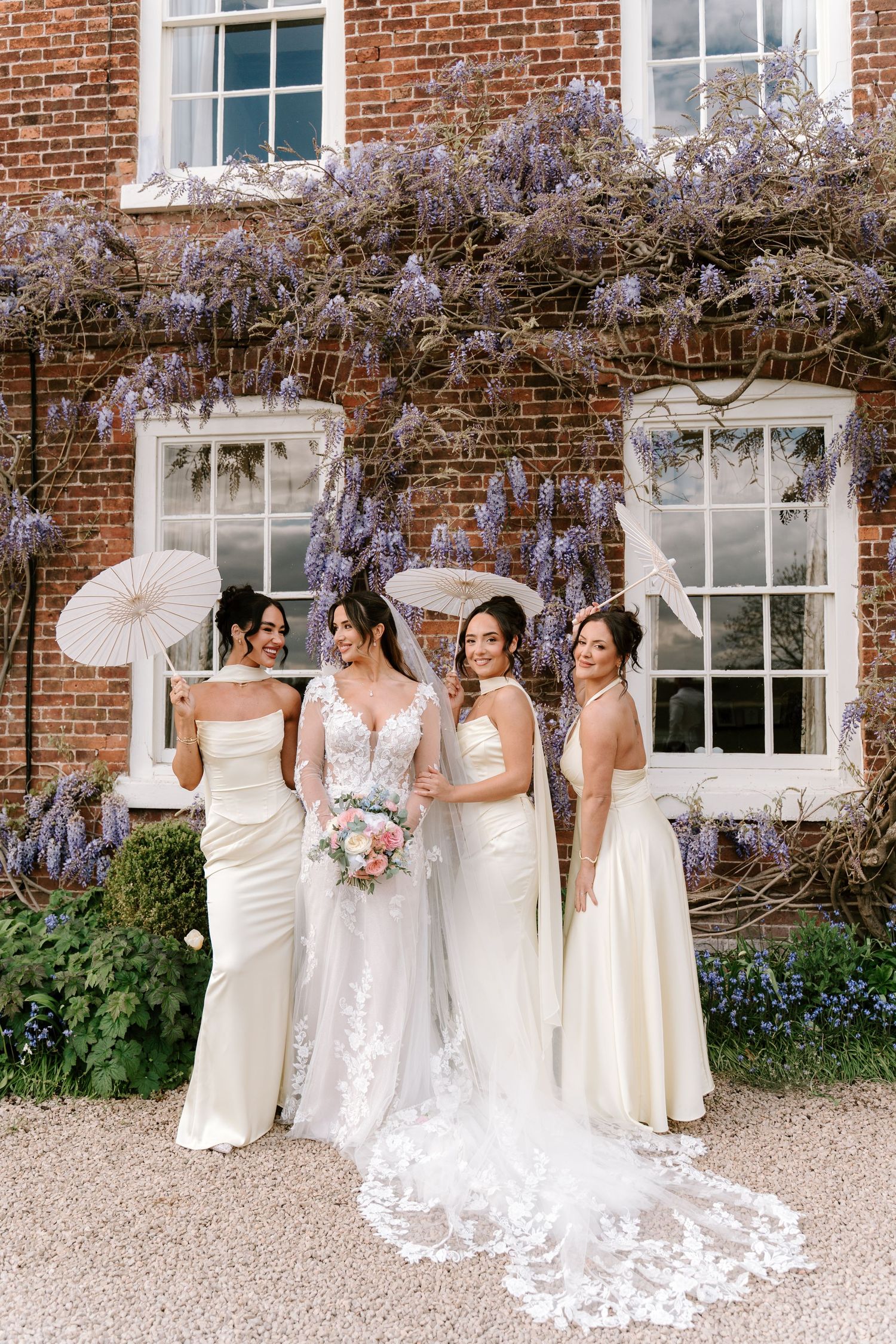 Bride and three bridesmaids in white gowns pose with parasols before a wisteria-covered brick manor house.