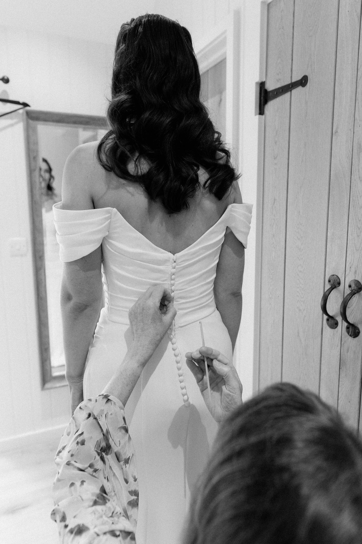 Bride in off-shoulder white gown having buttons fastened by bridesmaid in black and white photo.