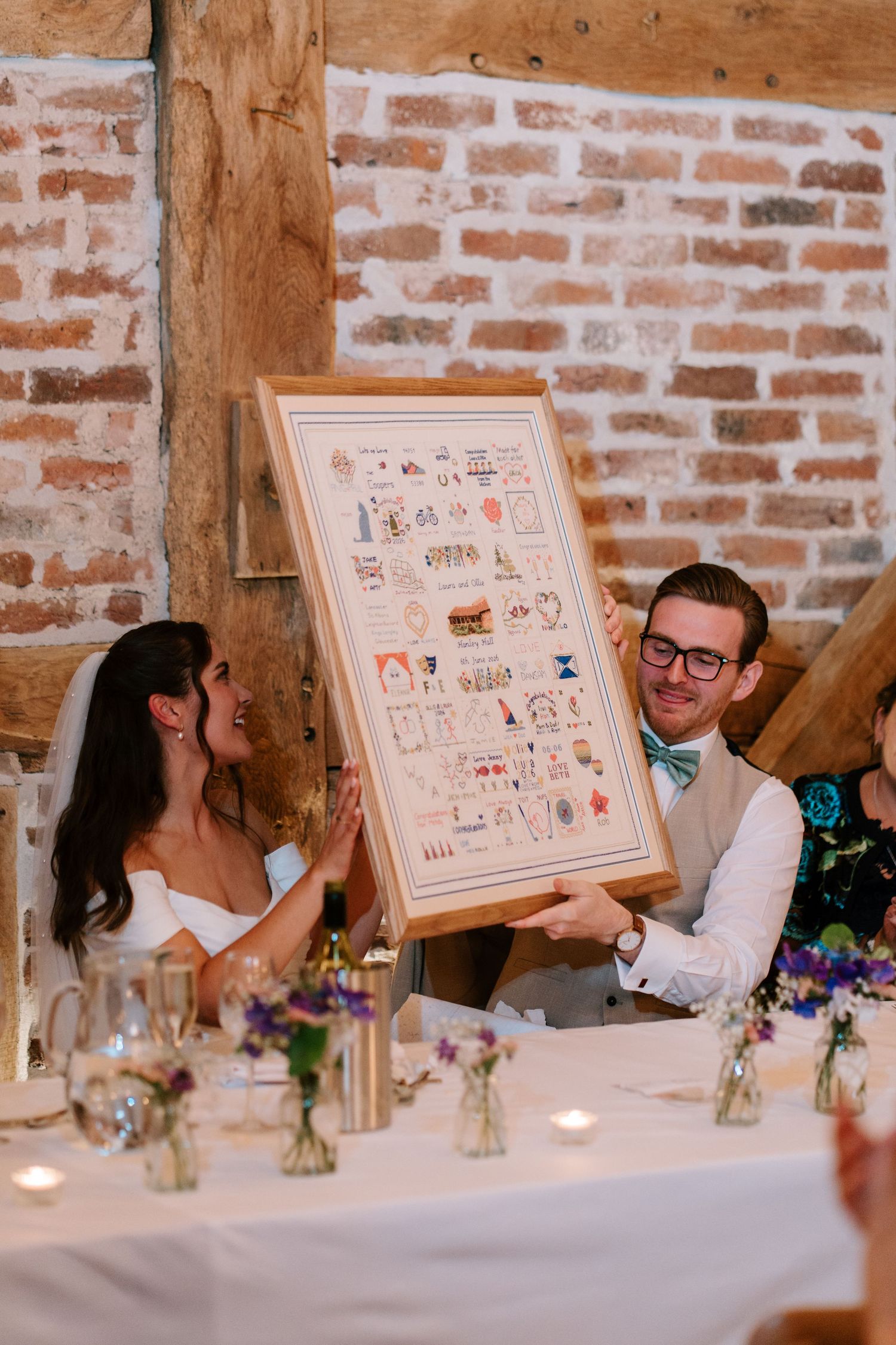 Bride and groom holding a framed cross-stitch artwork at their wedding reception in a rustic brick barn venue.