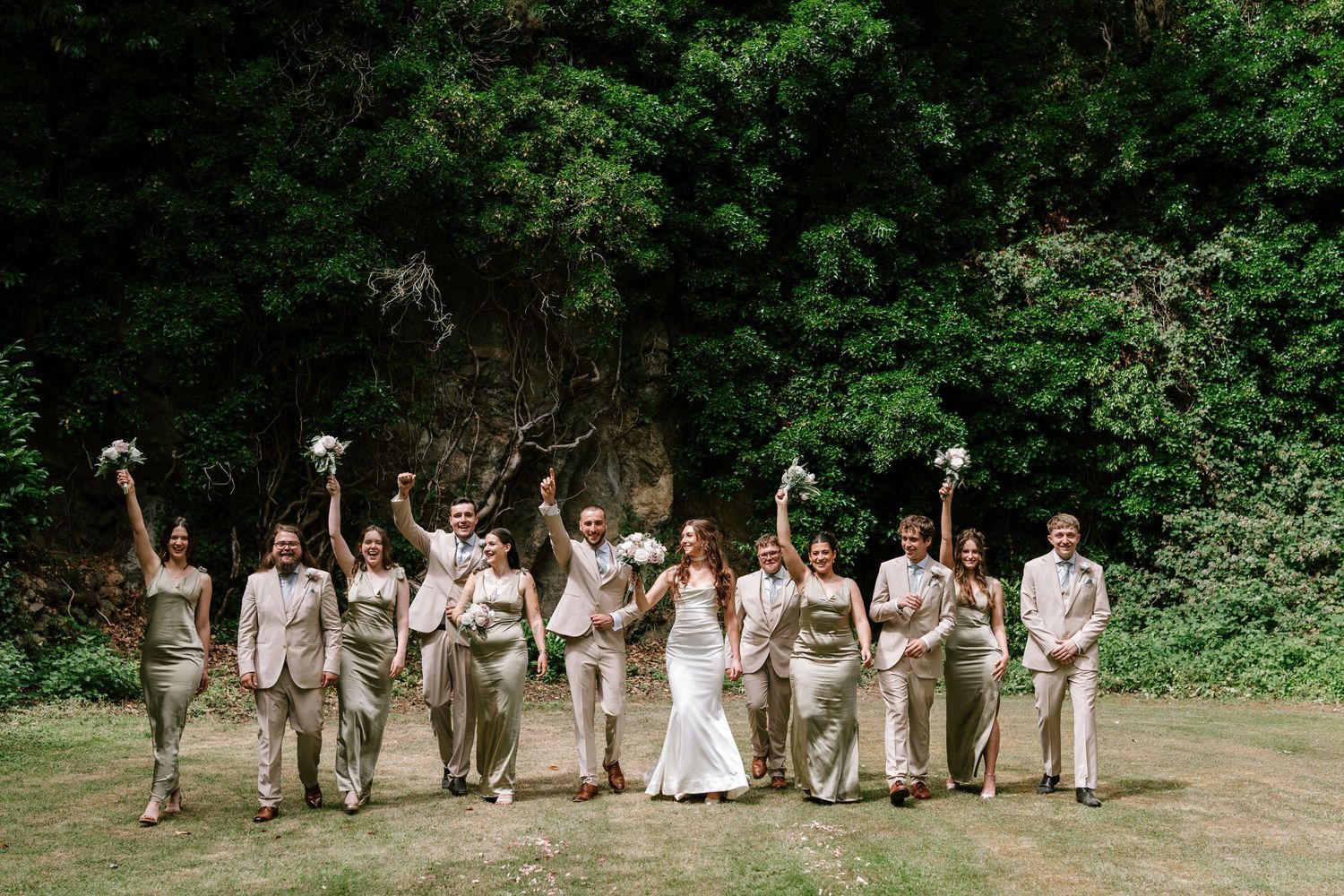 Joyful wedding party celebrates outdoors in sage and neutral attire surrounded by lush greenery.