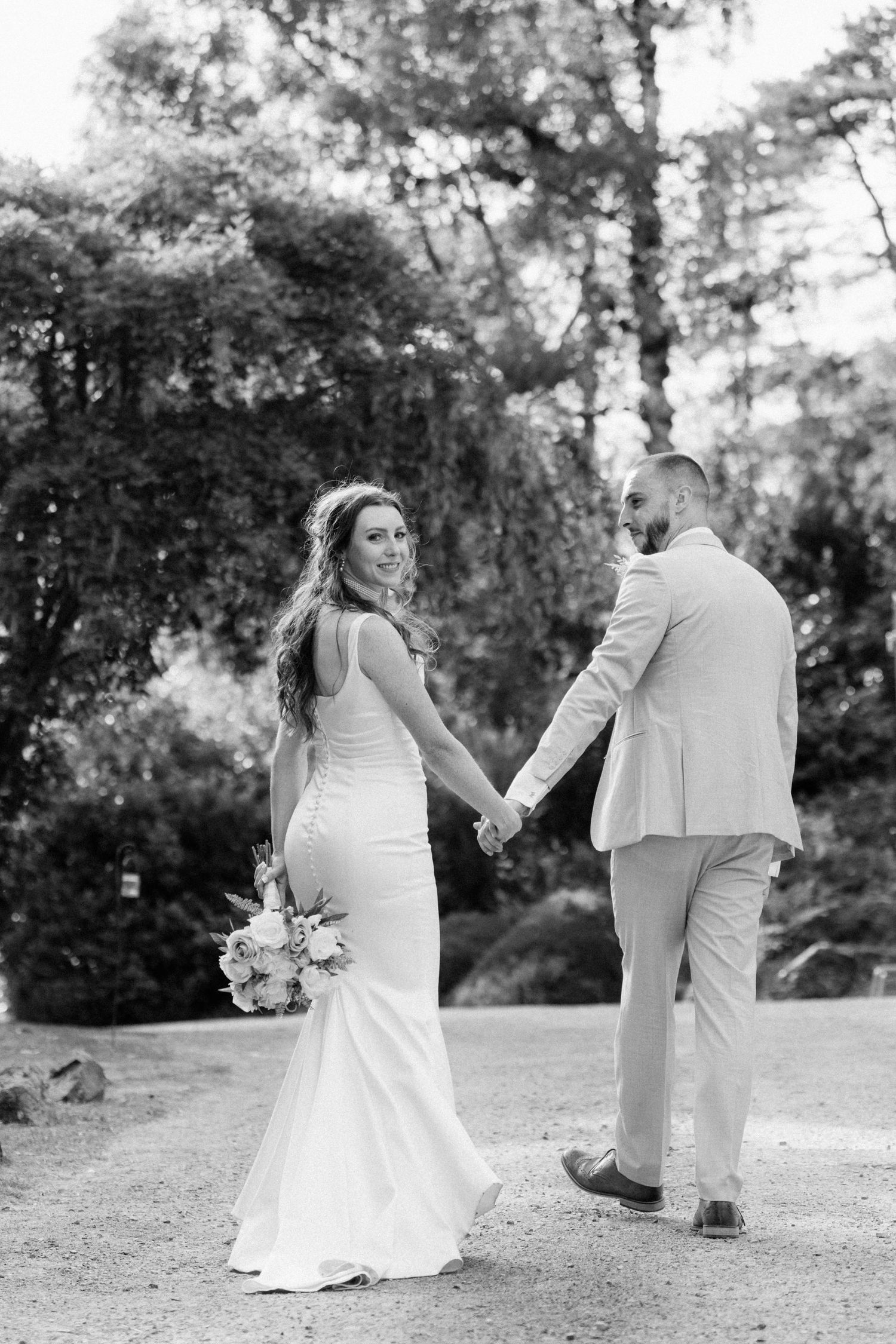 Black and white photo of a bride and groom holding hands, walking through a lush garden on their wedding day.