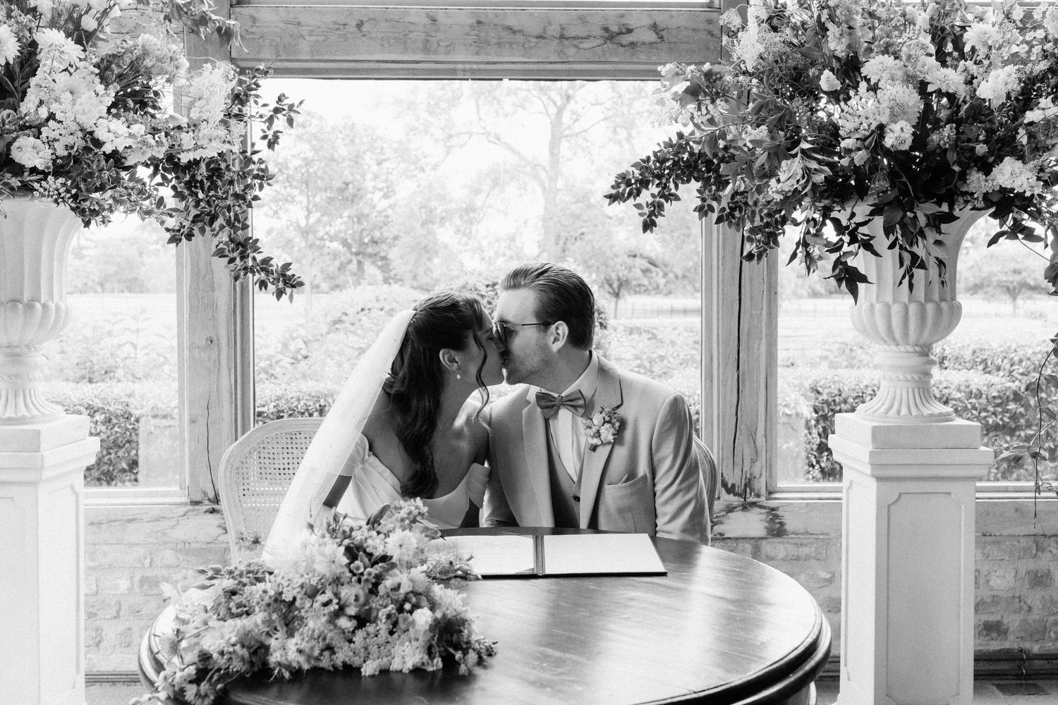 Black and white photo of a bride and groom sharing a kiss while signing their wedding register, surrounded by floral arrangements.
