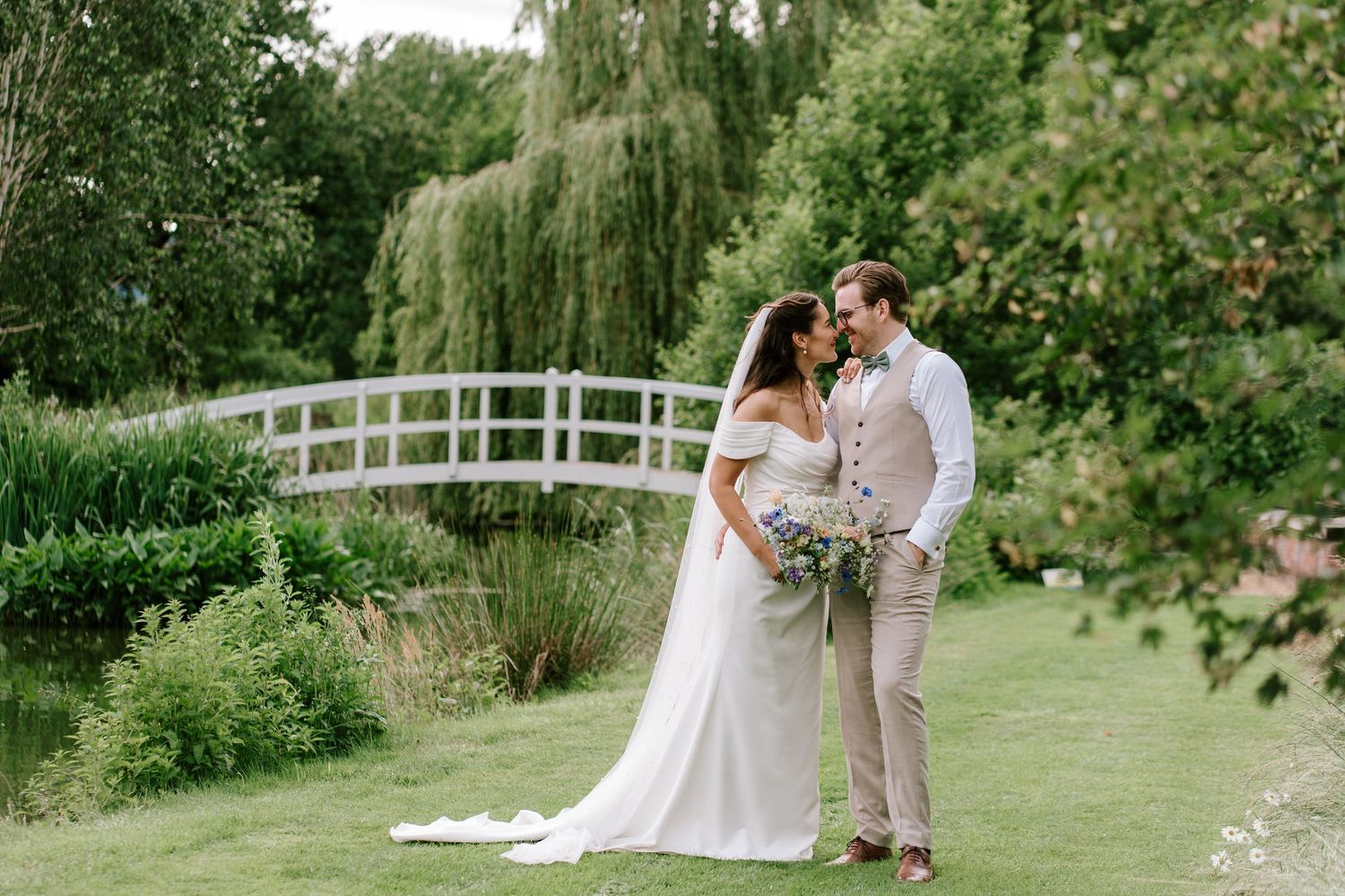 Bride and groom share romantic moment by white bridge in lush green garden with willow tree backdrop.