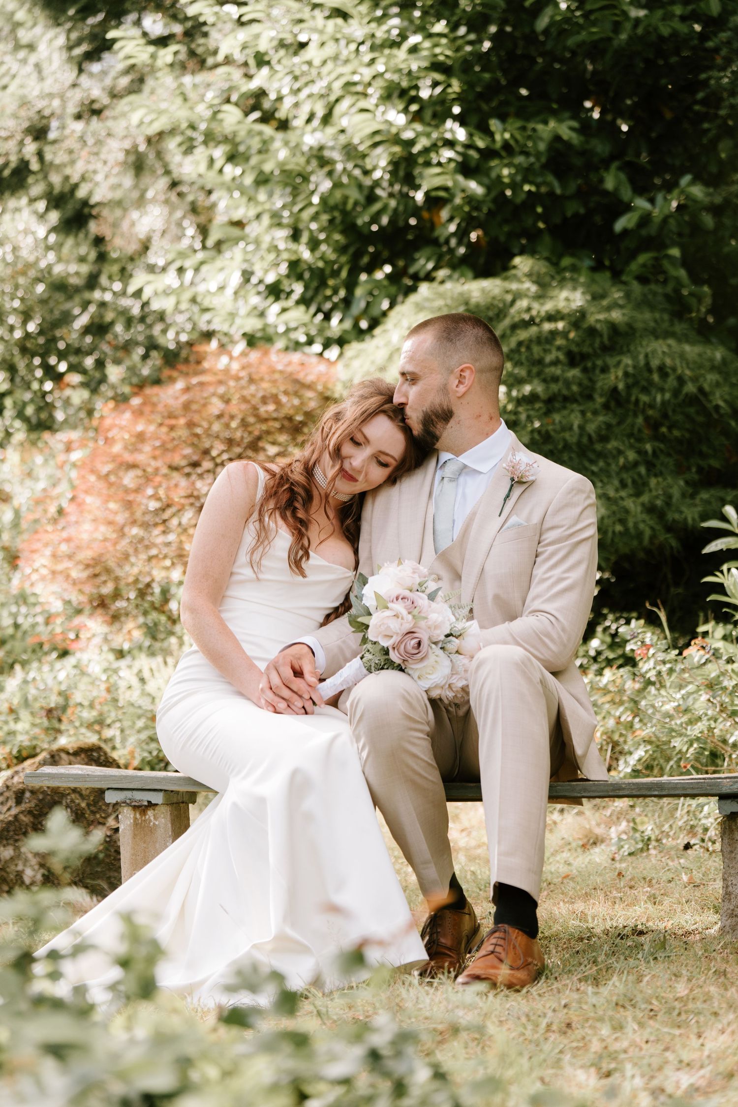Bride and groom sitting on a wooden bench in a lush garden, sharing a tender moment on their wedding day.