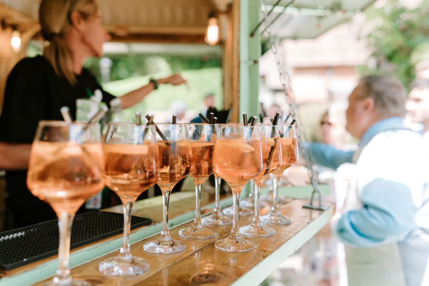 Rows of rose spritz cocktails in wine glasses lined up at an outdoor wedding bar with bartender serving guests.