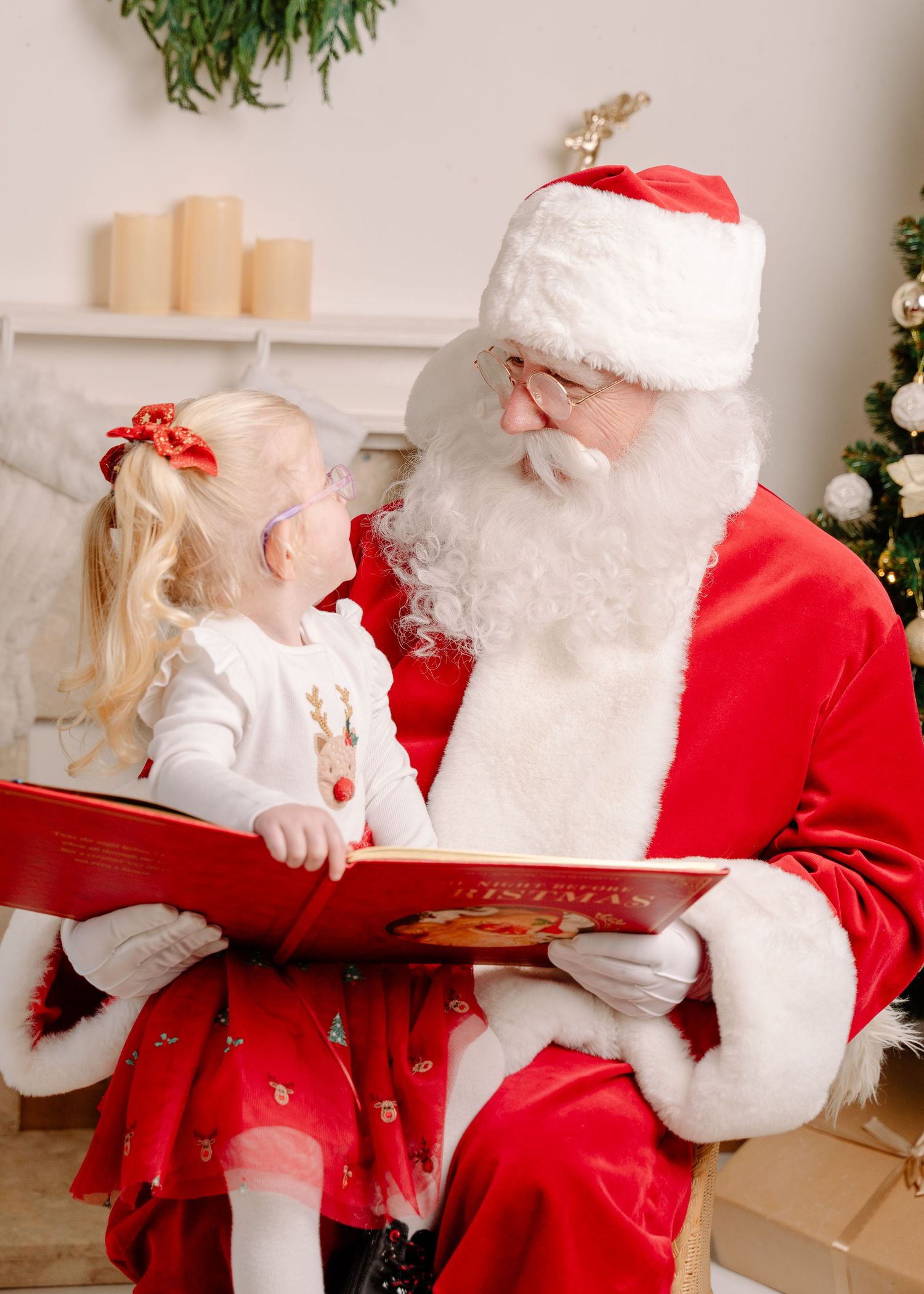 A young girl in a red skirt sits on Santa Claus's lap reading a Christmas book near a decorated tree.