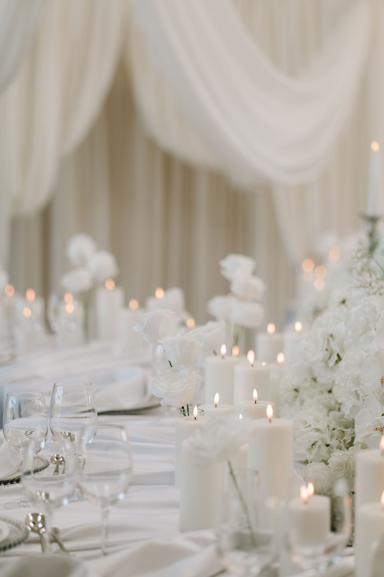 Elegant all-white wedding reception with candles, white floral arrangements, and draped fabric backdrop.