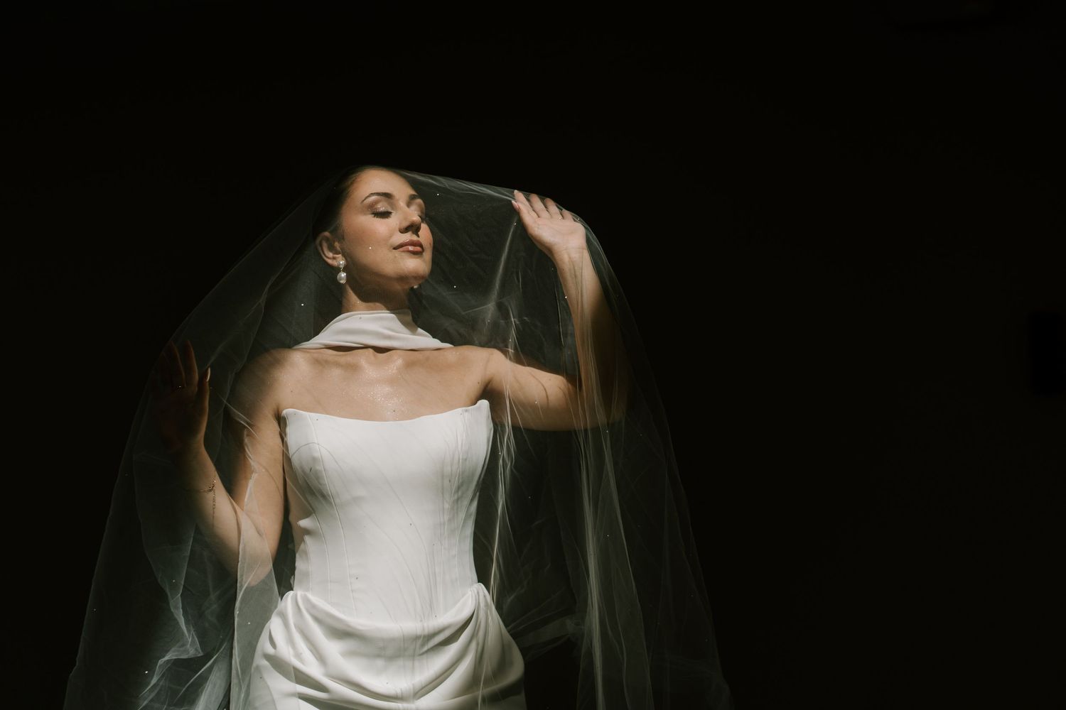 Bride in white strapless gown lifting sheer veil against dark background, elegant wedding portrait photography.