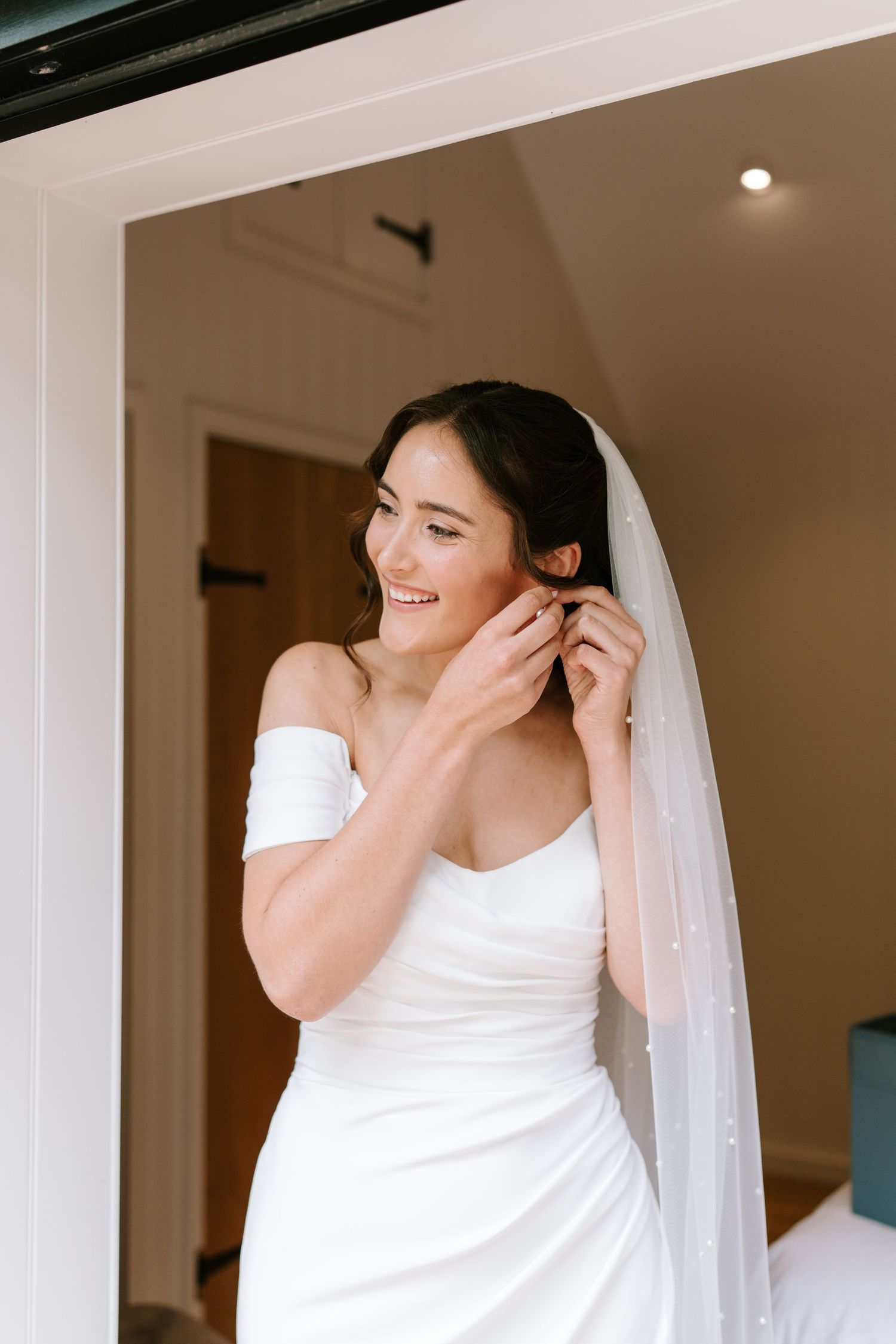 Smiling bride in off-shoulder white gown and veil putting on earrings while getting ready on her wedding day.
