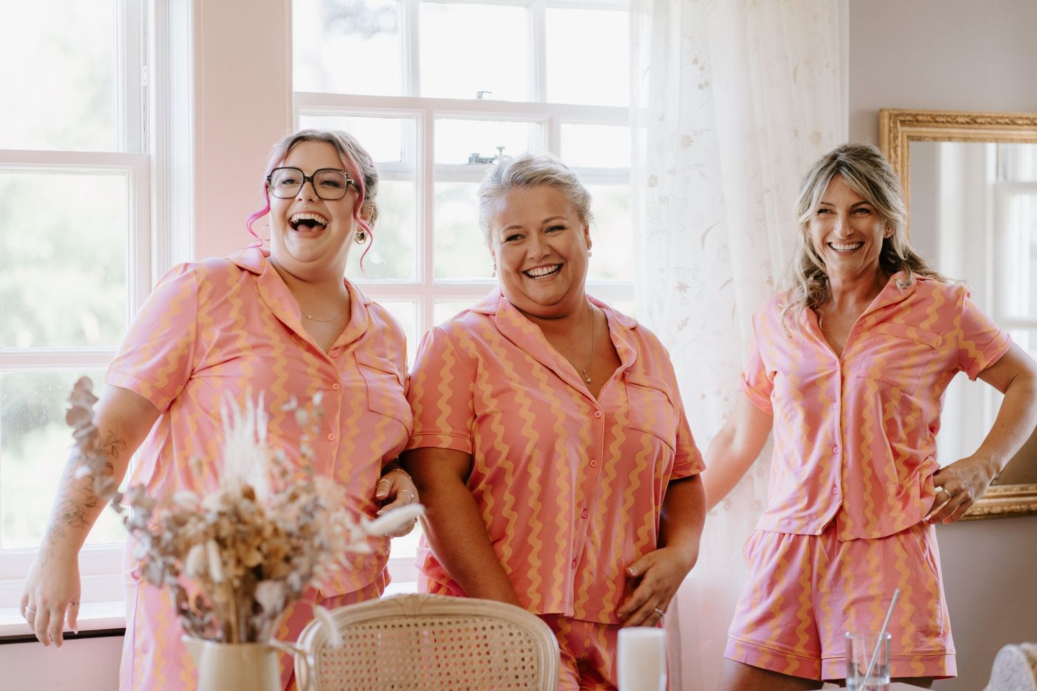 Three women laughing in matching pink pajama sets standing together in a bright, elegant room.