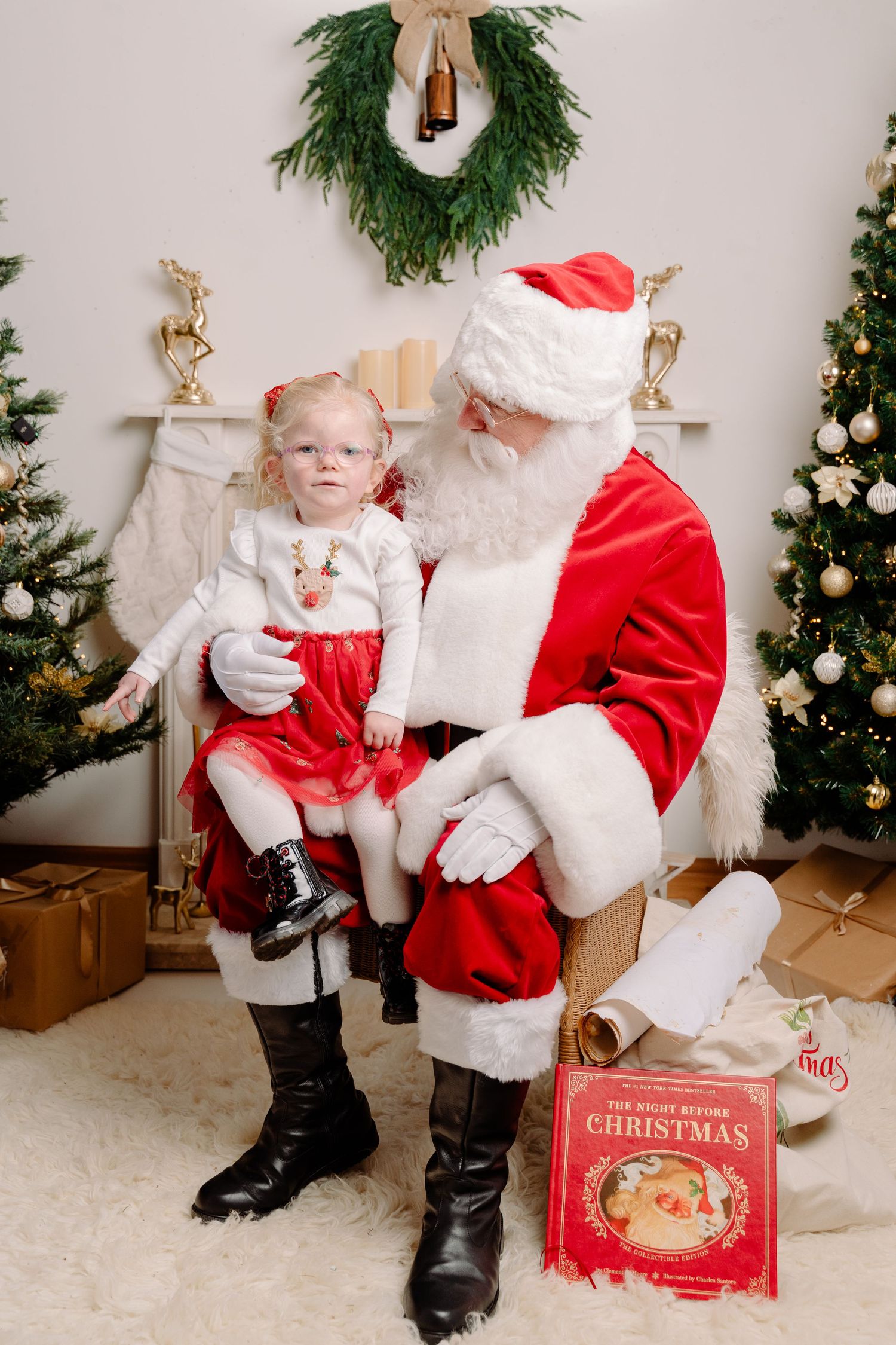 A toddler girl in festive outfit sits on Santa Claus lap in a decorated Christmas holiday setting.