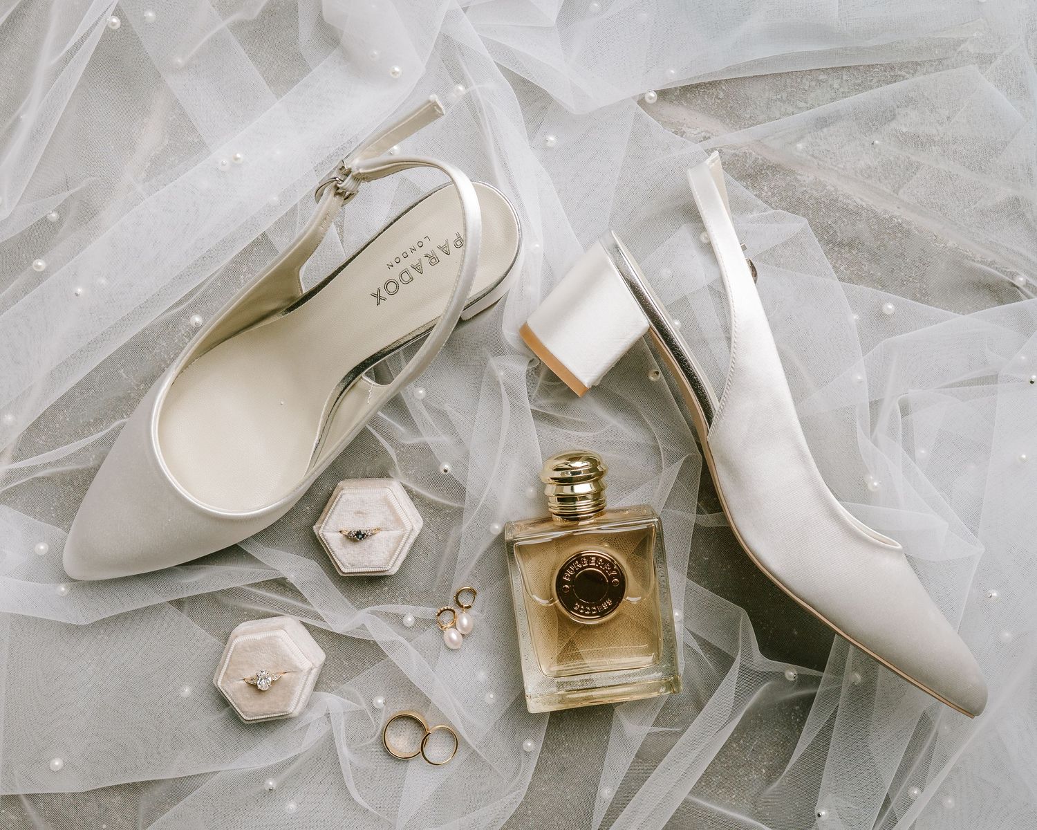 Elegant bridal flat lay featuring white slingback heels, gold perfume bottle, ring box, and pearl earrings on sheer tulle fabric.