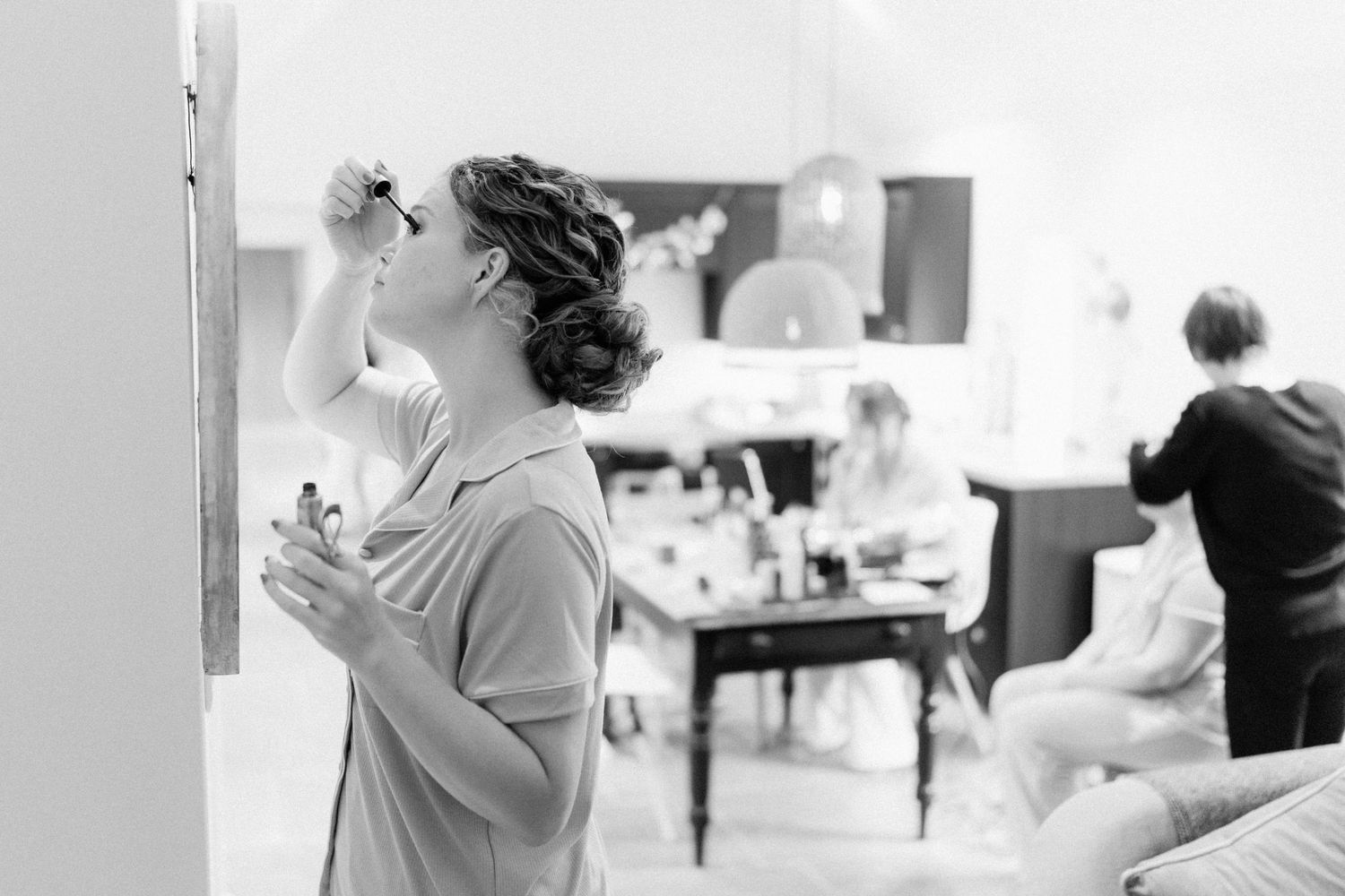 Black and white photo of a woman applying makeup while others prepare in a bright, elegant room.