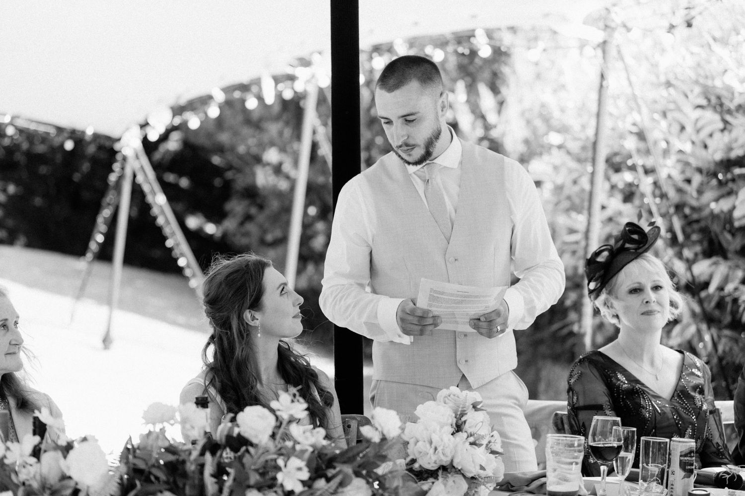 Groom delivering heartfelt speech at wedding reception head table with floral arrangements in black and white.