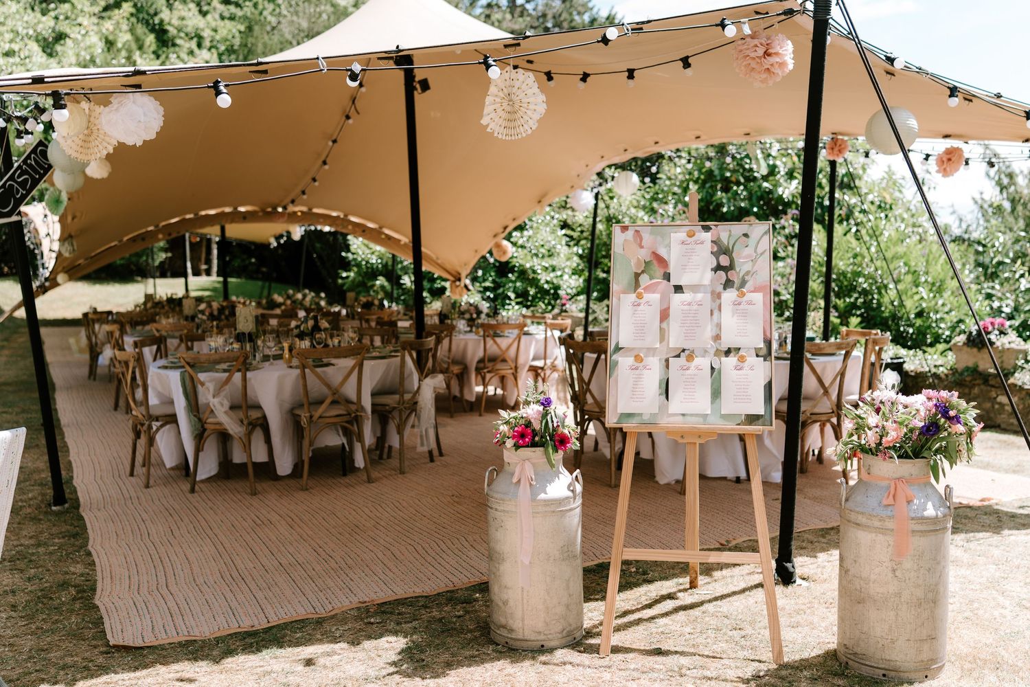 Outdoor wedding reception under stretch tent with fairy lights, round tables, and floral seating chart display.