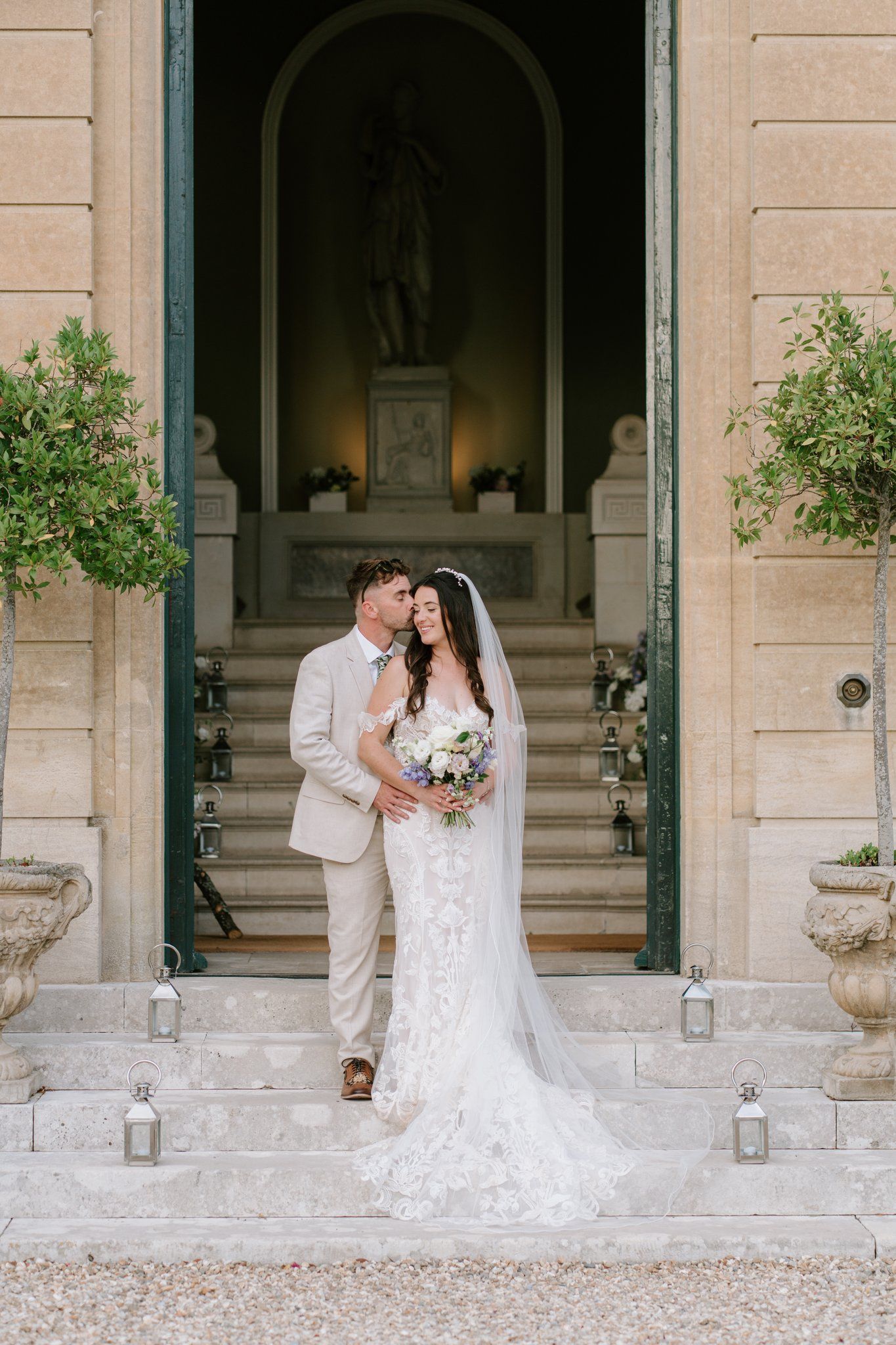Bride and groom pose on stone steps of elegant villa, she holds floral bouquet in lace gown with veil, he wears beige suit.