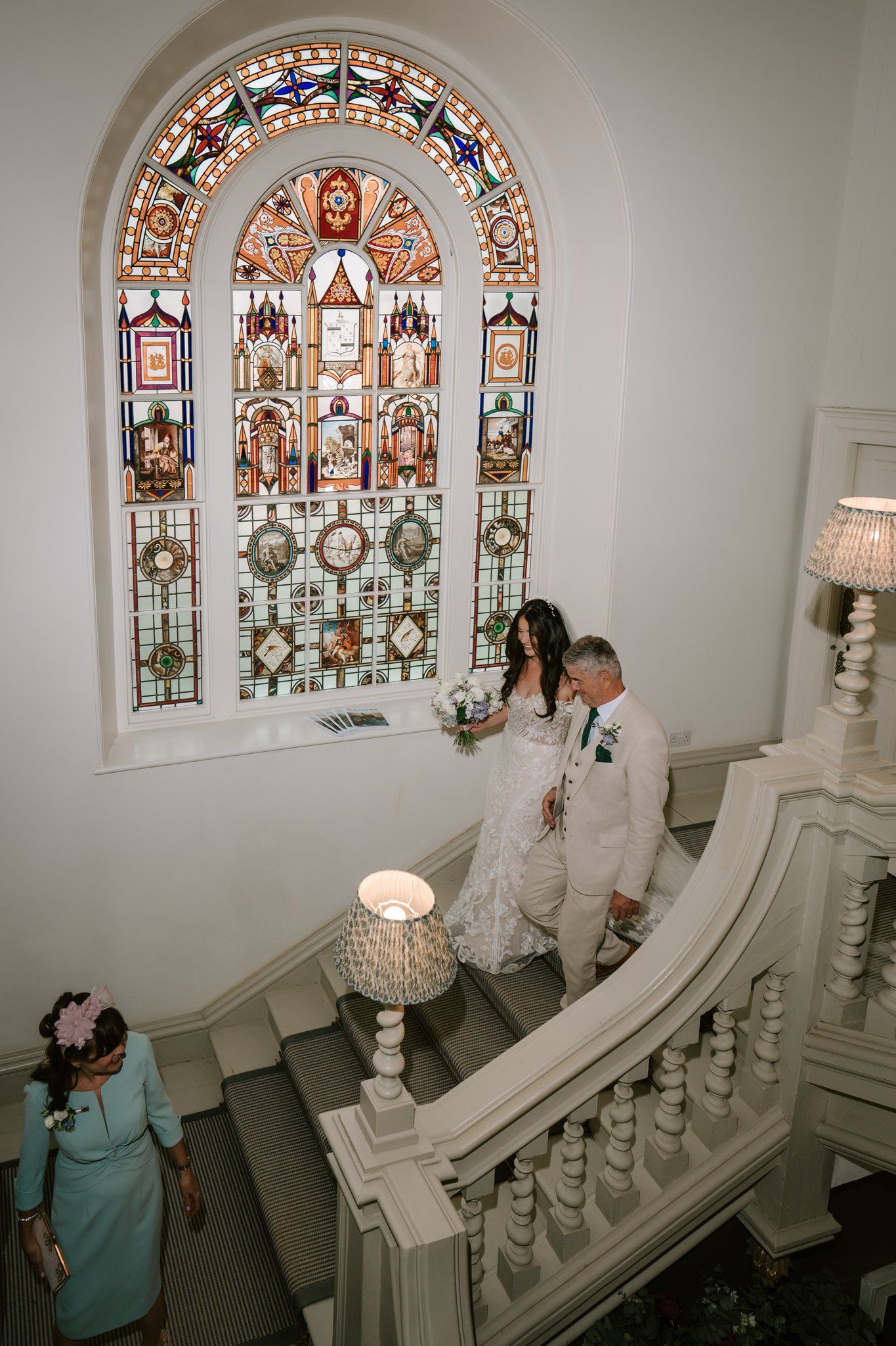 Bride and groom descend elegant white staircase beneath colorful stained glass window in historic wedding venue.