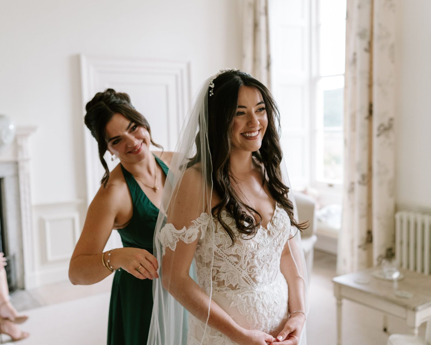 Bride in lace wedding dress and veil smiles as bridesmaid in green dress helps with final preparations.