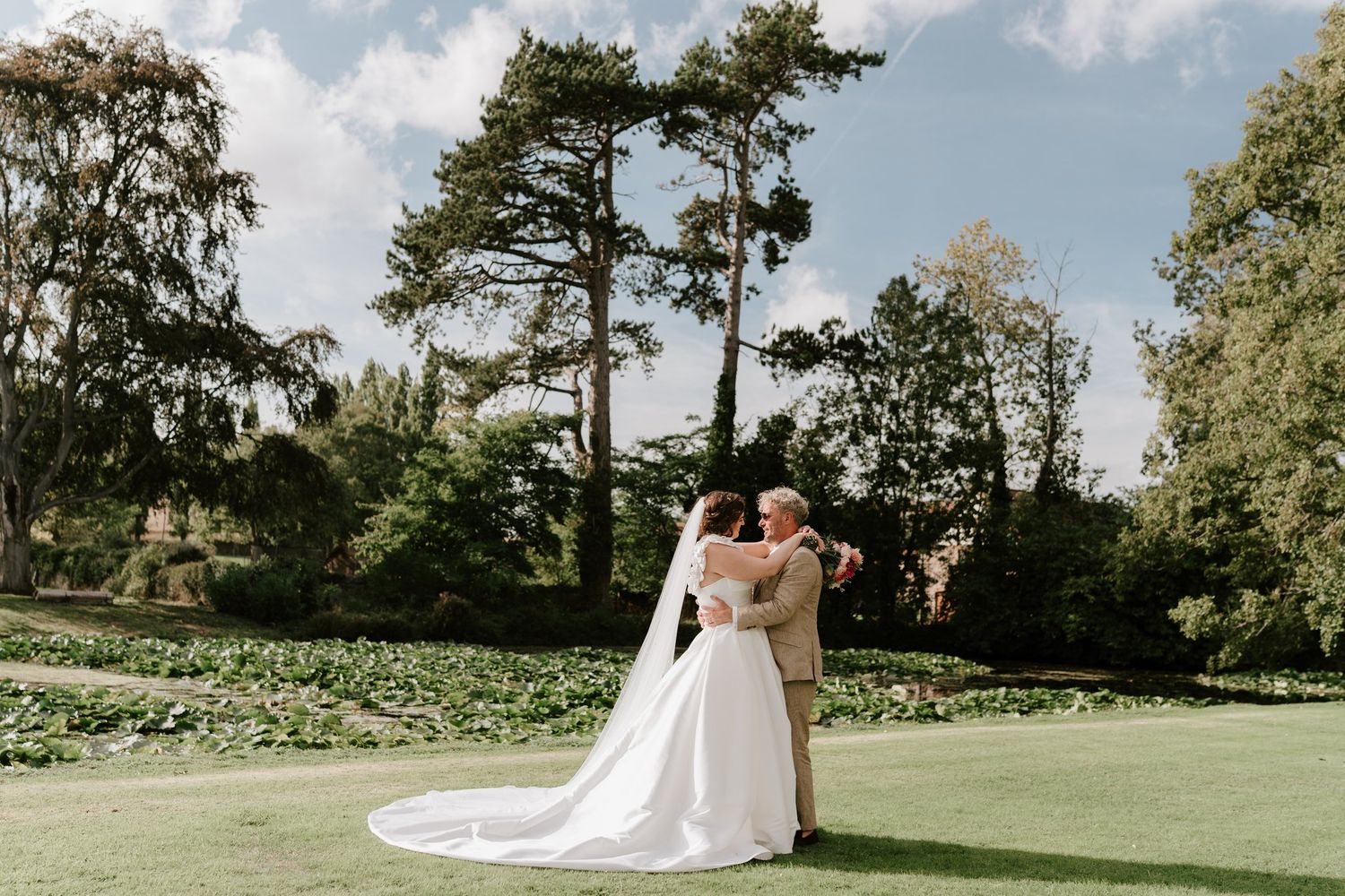 Bride and groom embracing on the lawn beneath tall trees at Brinsop Court