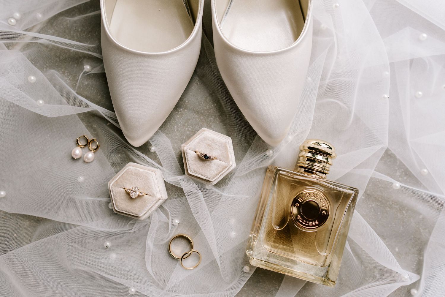 Elegant bridal flat lay with white heels, perfume bottle, gold rings, pearl earrings, and a sheer veil on marble.
