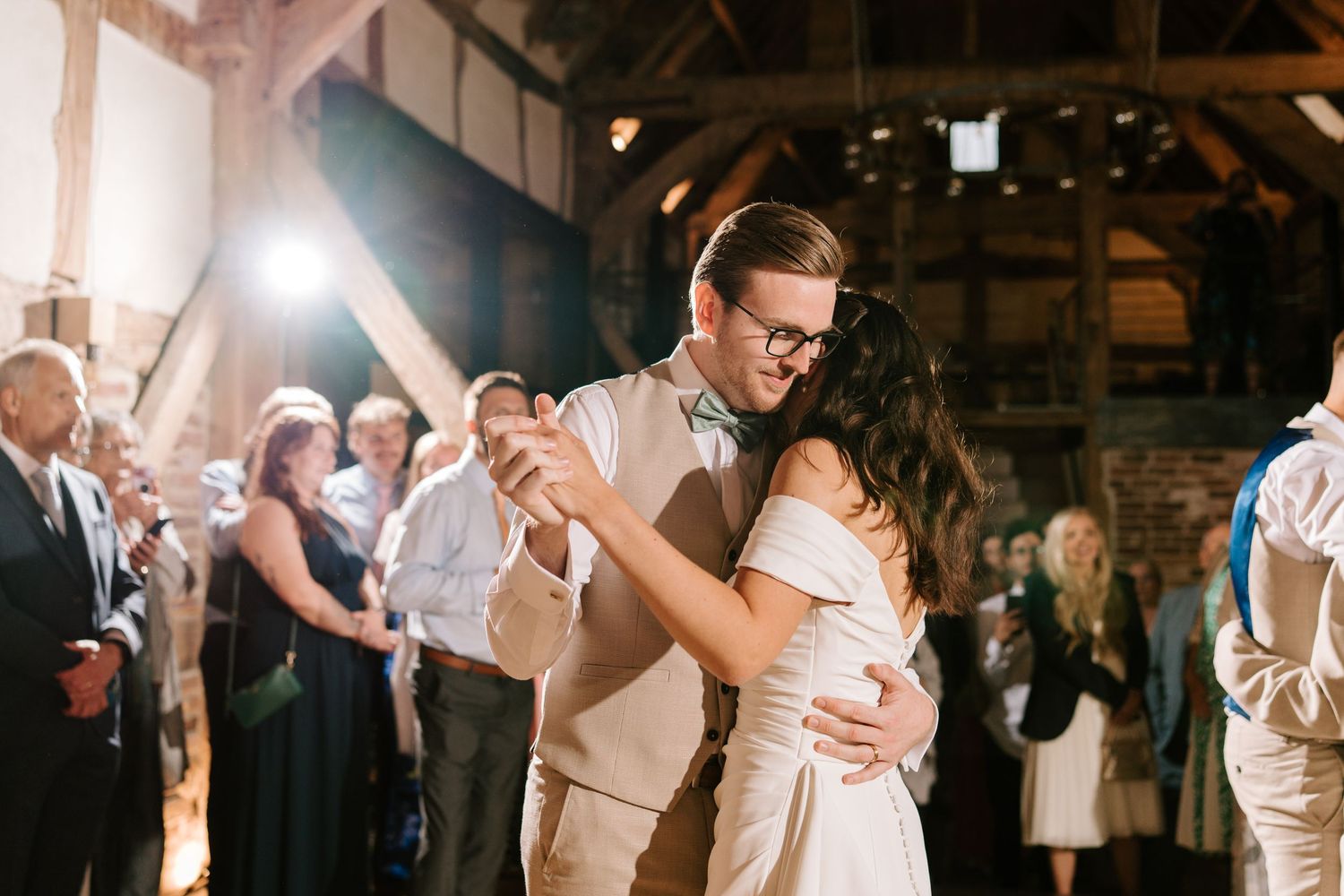 Bride and groom share their first dance in a warmly lit rustic barn surrounded by wedding guests.