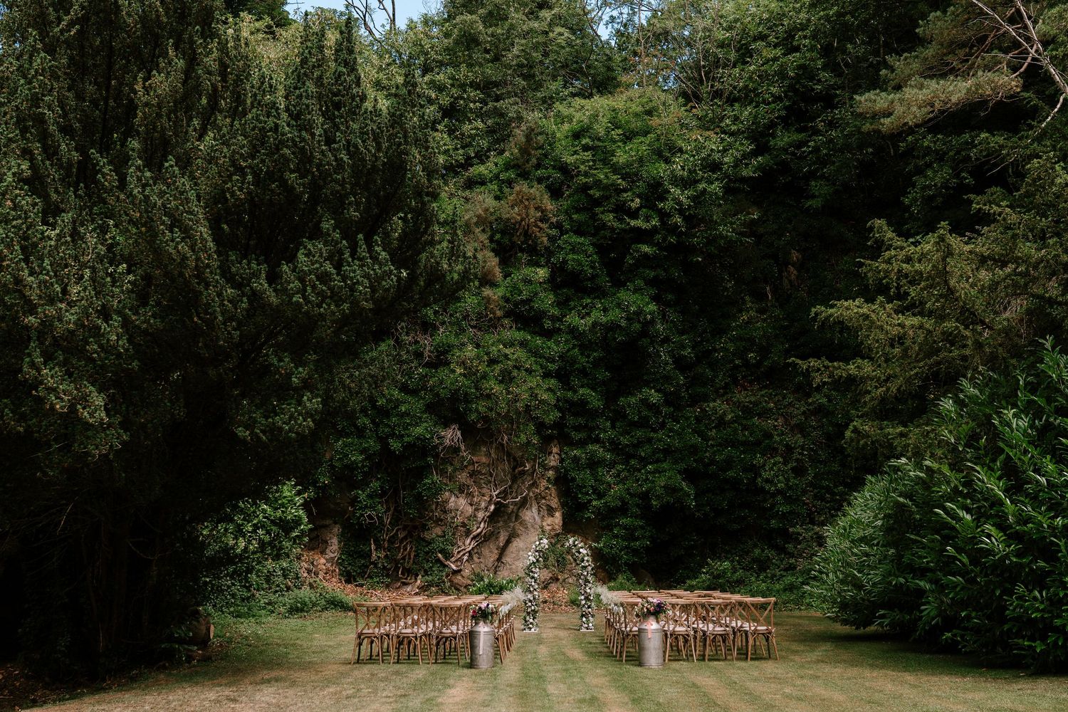 Elegant outdoor wedding ceremony venue with wooden chairs on green lawn surrounded by tall trees.