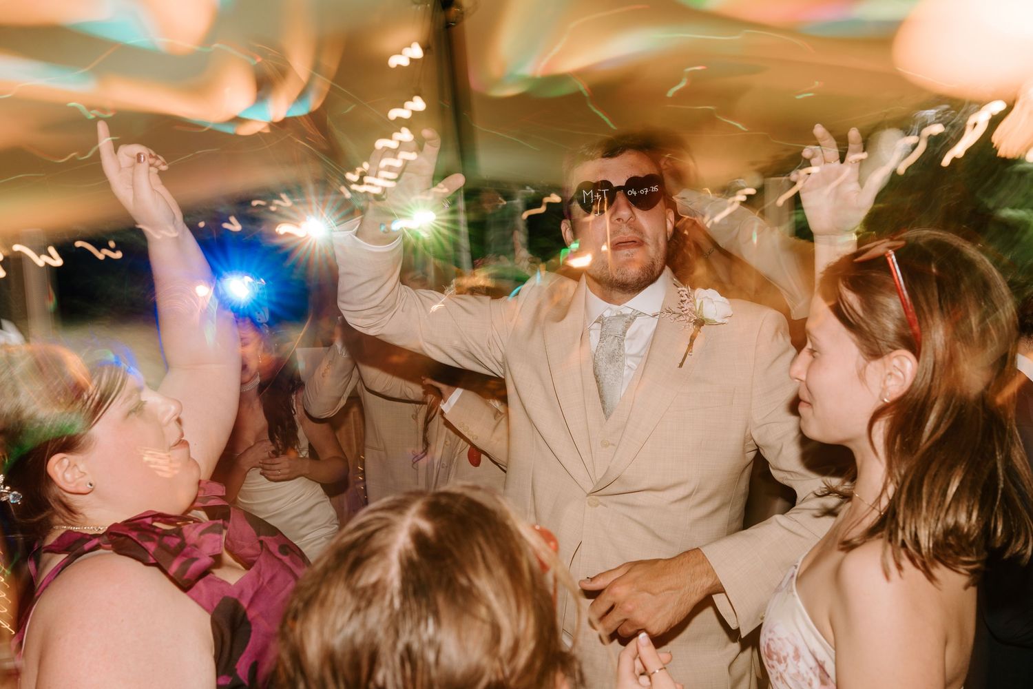 Groom in tan suit dancing with sparklers and sunglasses surrounded by guests at lively wedding reception.