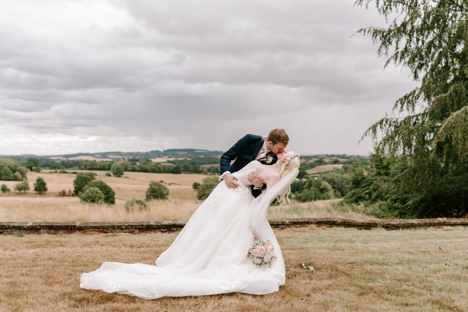 Groom dips bride in white gown outdoors with rolling countryside backdrop on their wedding day.