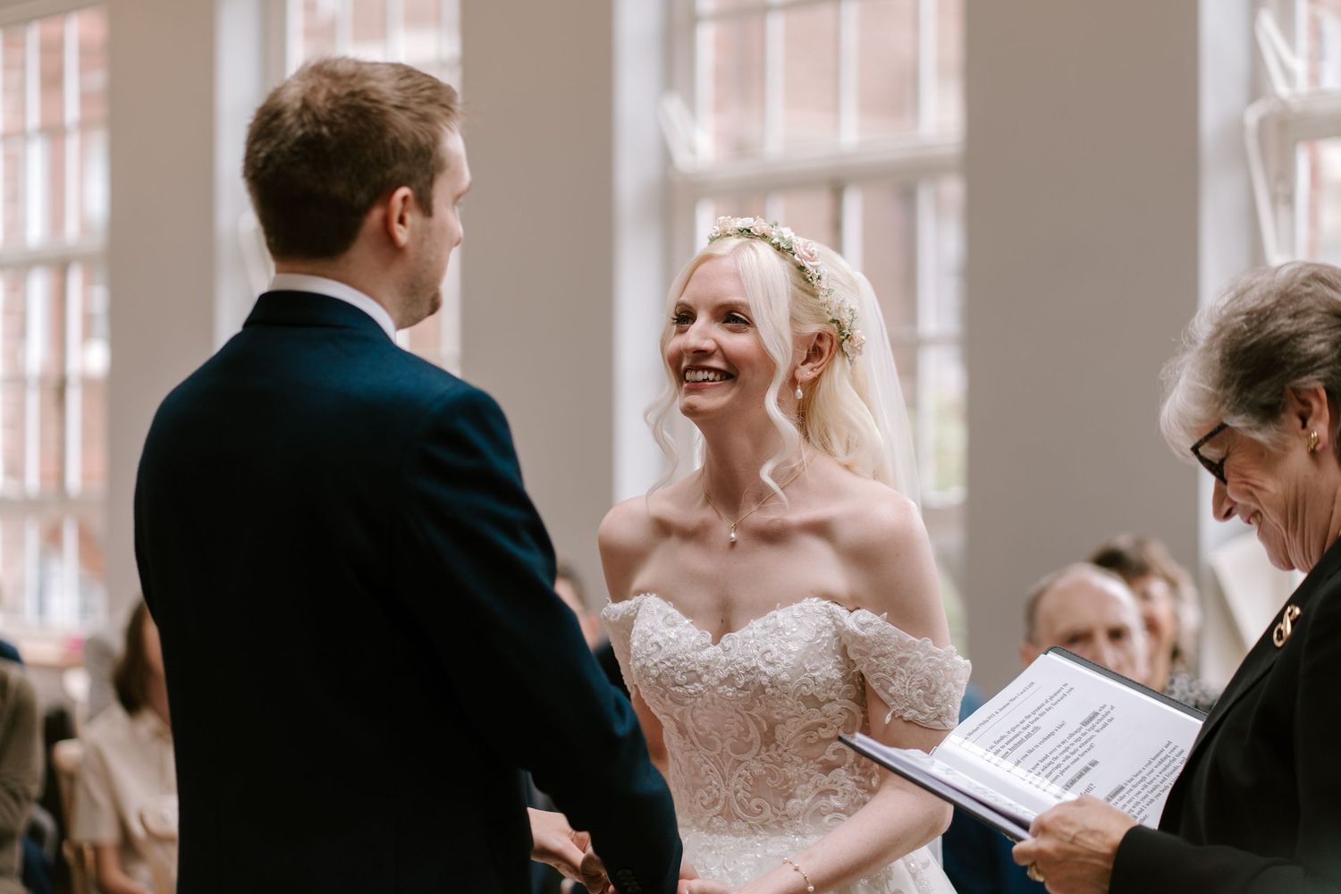 Bride smiling joyfully at groom during wedding ceremony, officiant holding script, guests in background.