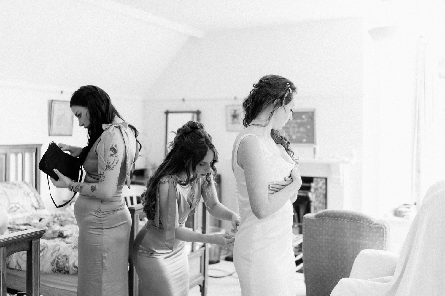 Black and white photo of bride being helped into wedding dress by bridesmaids in a bright white room.