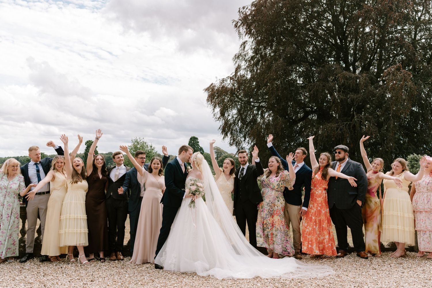 Wedding party celebrating outdoors, bride and groom kissing while guests cheer with arms raised under cloudy sky.