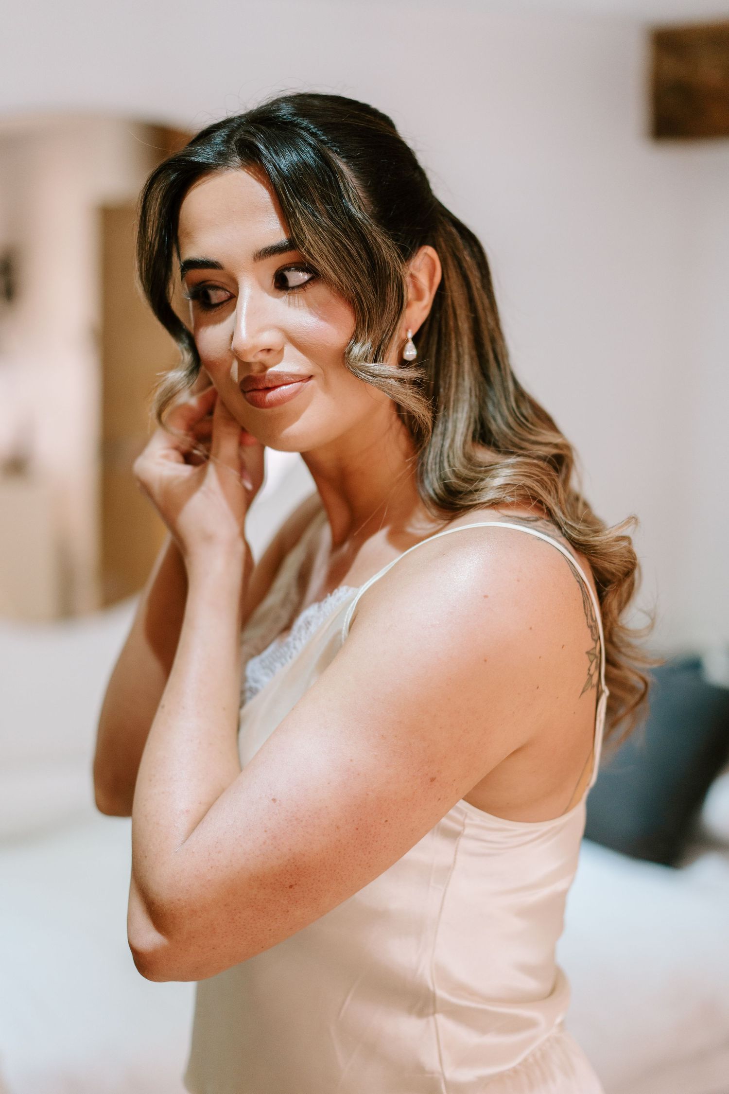 Bride putting on earrings in a champagne slip dress, with soft curled hair and elegant makeup.
