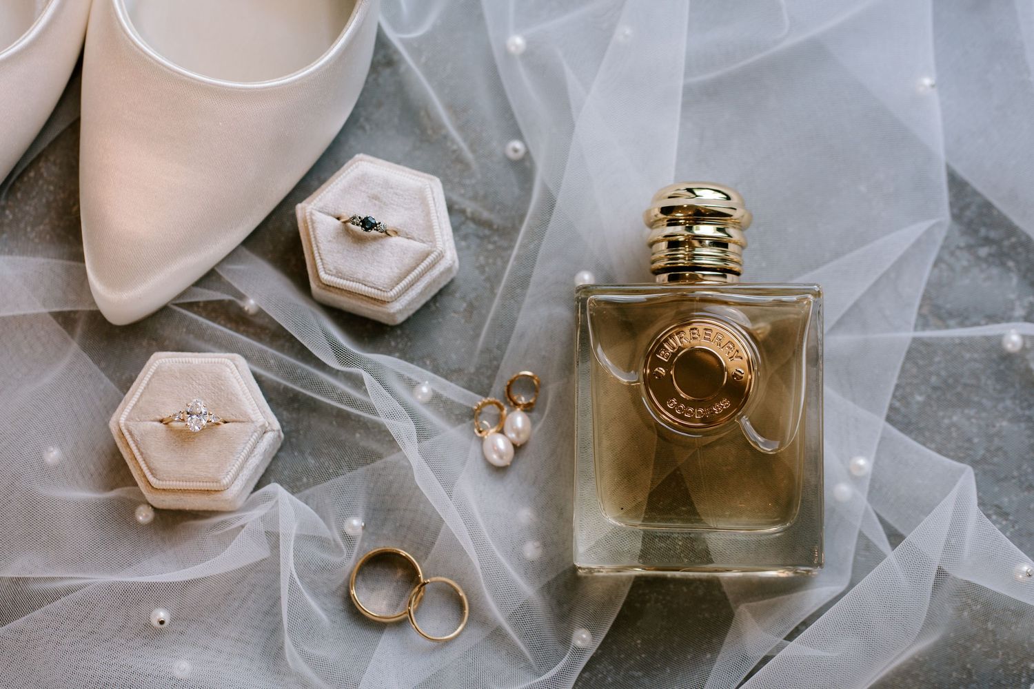 Elegant bridal flat lay with perfume, diamond rings, pearl earrings, gold bands, and white heels on sheer veil.