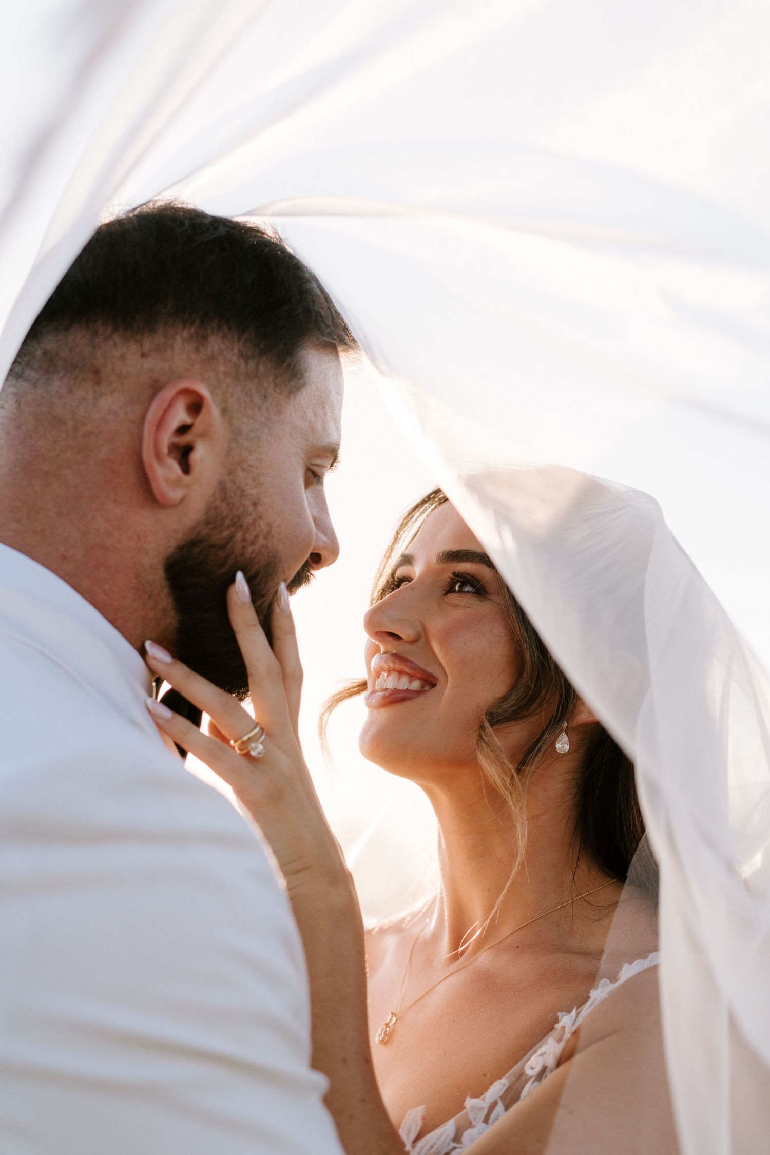 Bride and groom share a romantic moment under a flowing white veil, smiling at each other on their wedding day.
