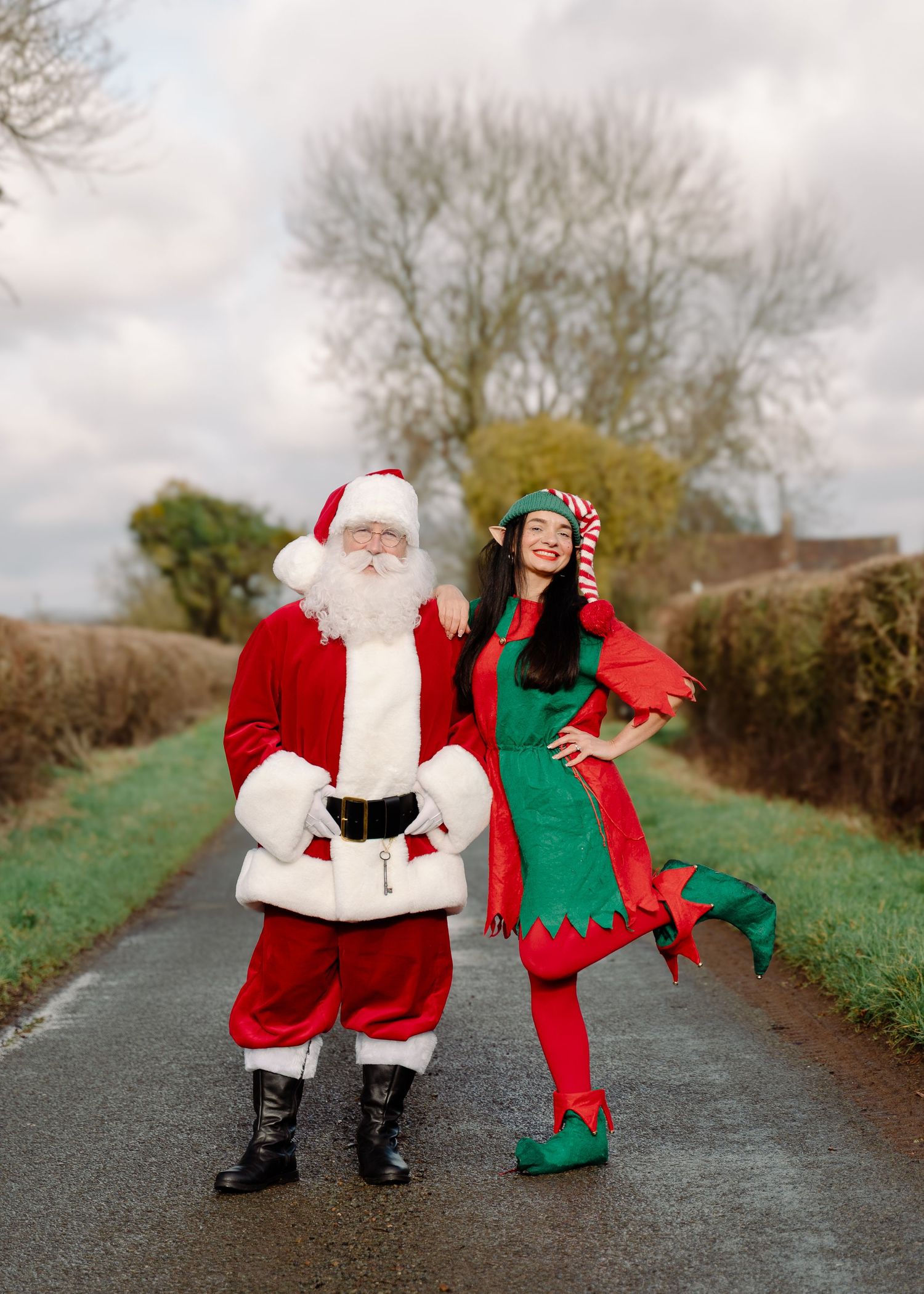 Santa Claus and a female Christmas elf in festive costumes pose together on a rural road with bare trees in winter.