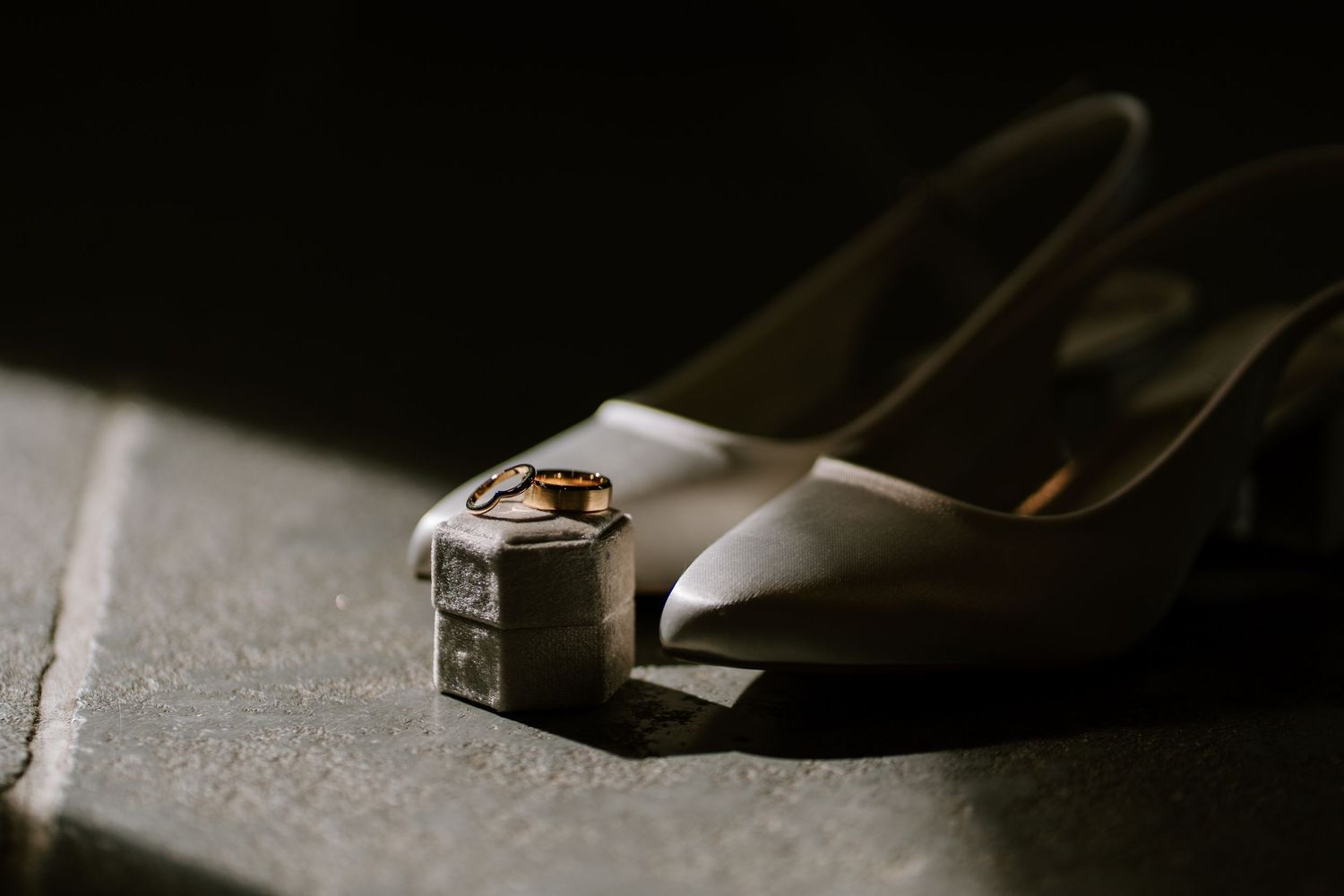 Wedding rings resting on a small silver box beside elegant ivory bridal heels on a stone surface in soft light.