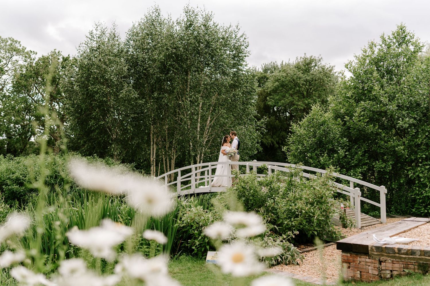 Newlywed couple embracing on a rustic white bridge surrounded by lush greenery and white daisies in a garden.