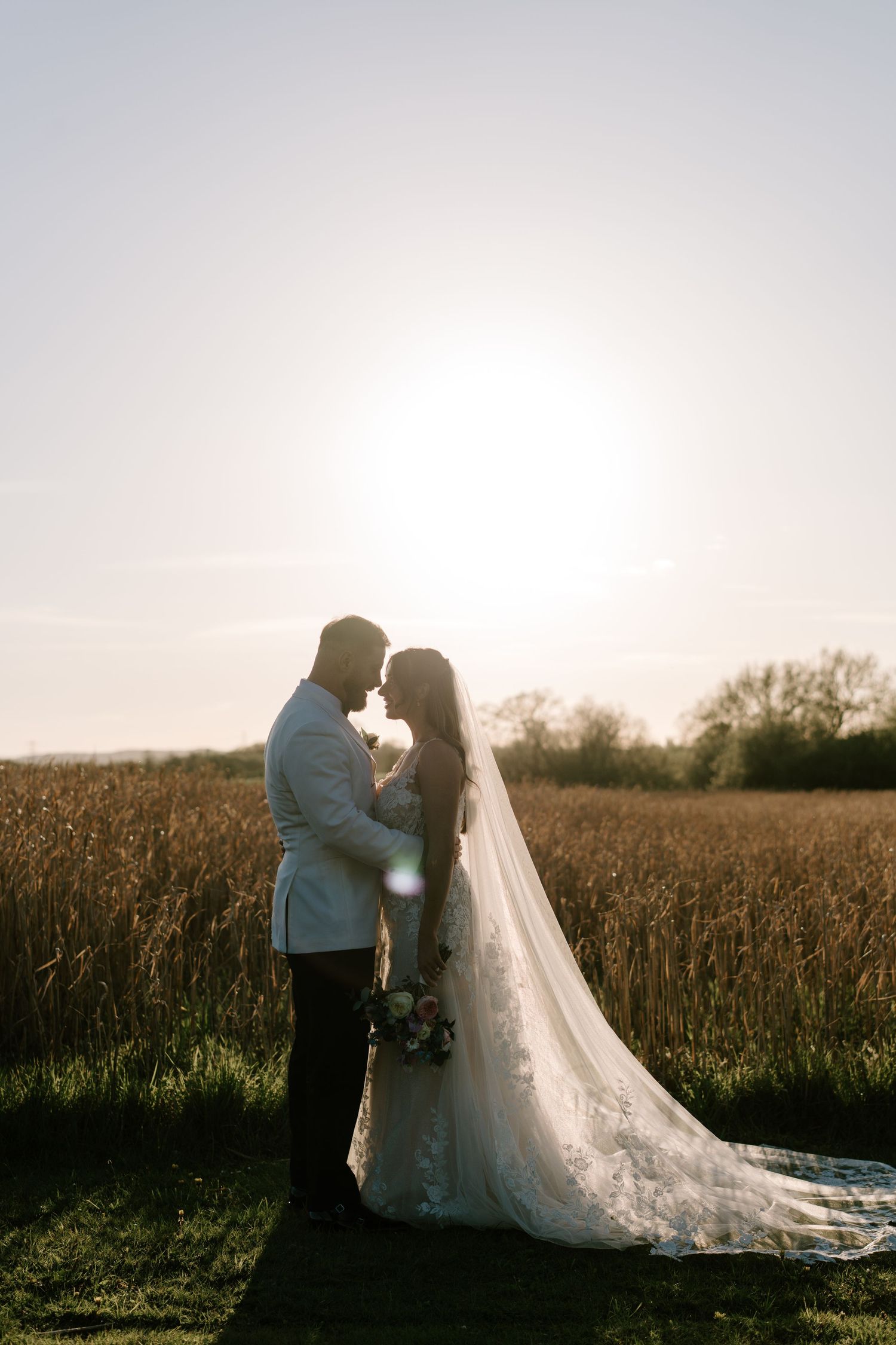 Bride and groom share a romantic sunset moment in a golden field, her long veil trailing gracefully behind her.