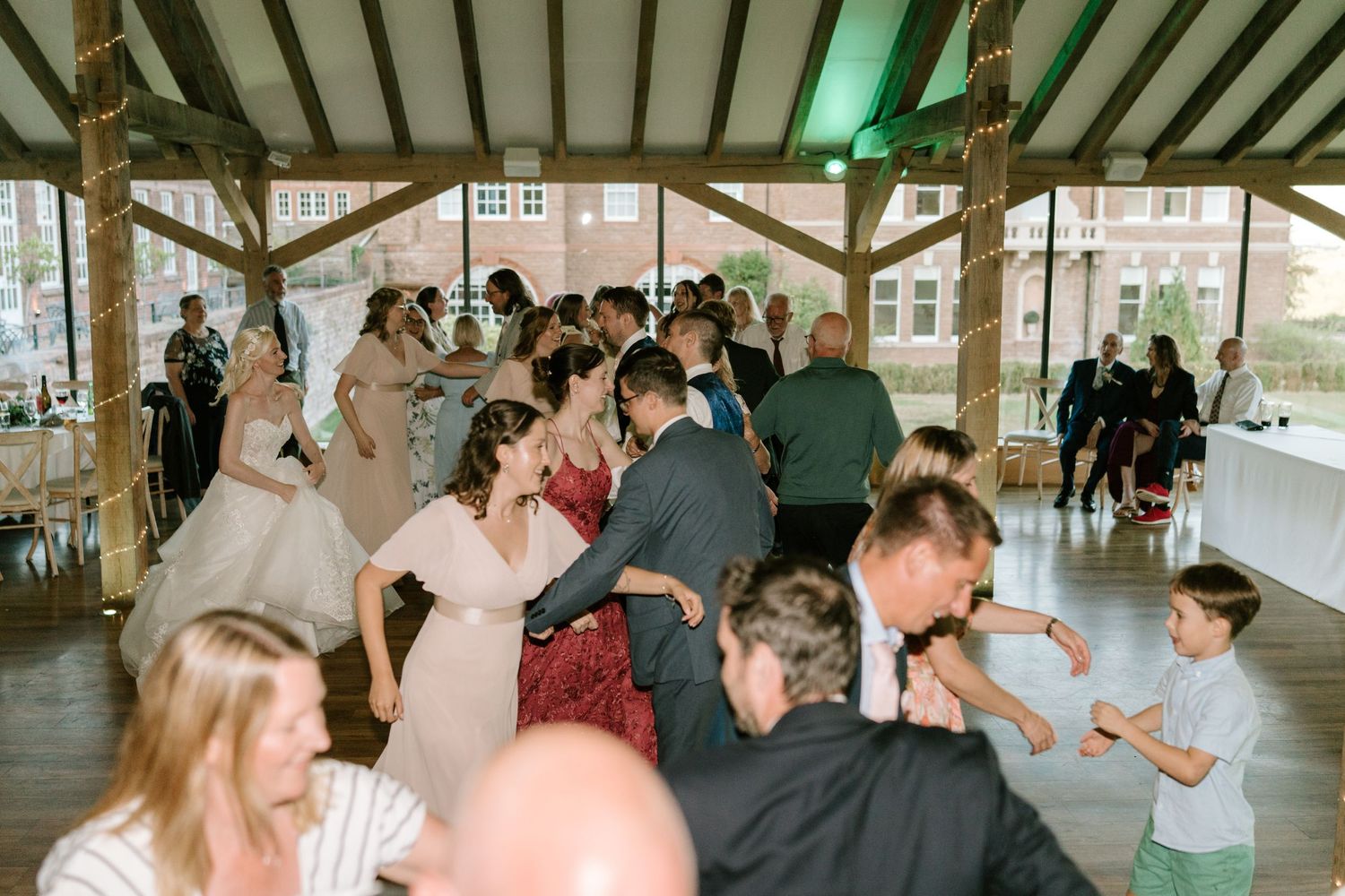 Guests dancing joyfully at an indoor wedding reception under a wooden pavilion with green accent lighting.