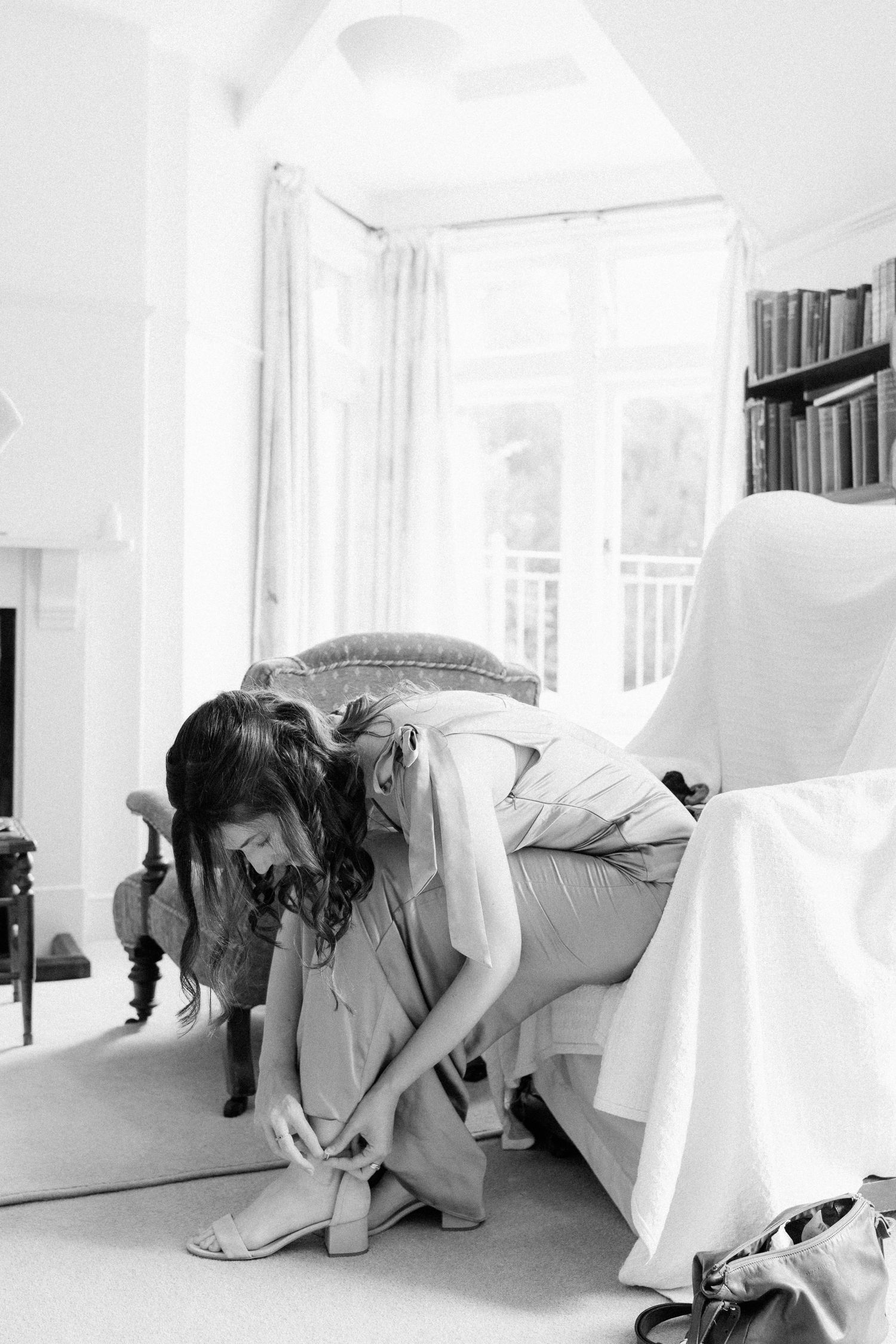 Bride sitting on bed putting on heeled sandals in bright white room while getting ready for wedding.