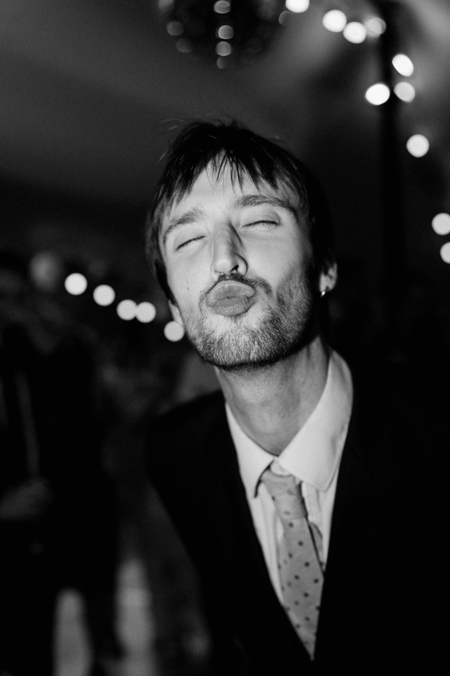 Black and white photo of a man in a suit making a kissing face at a wedding reception.