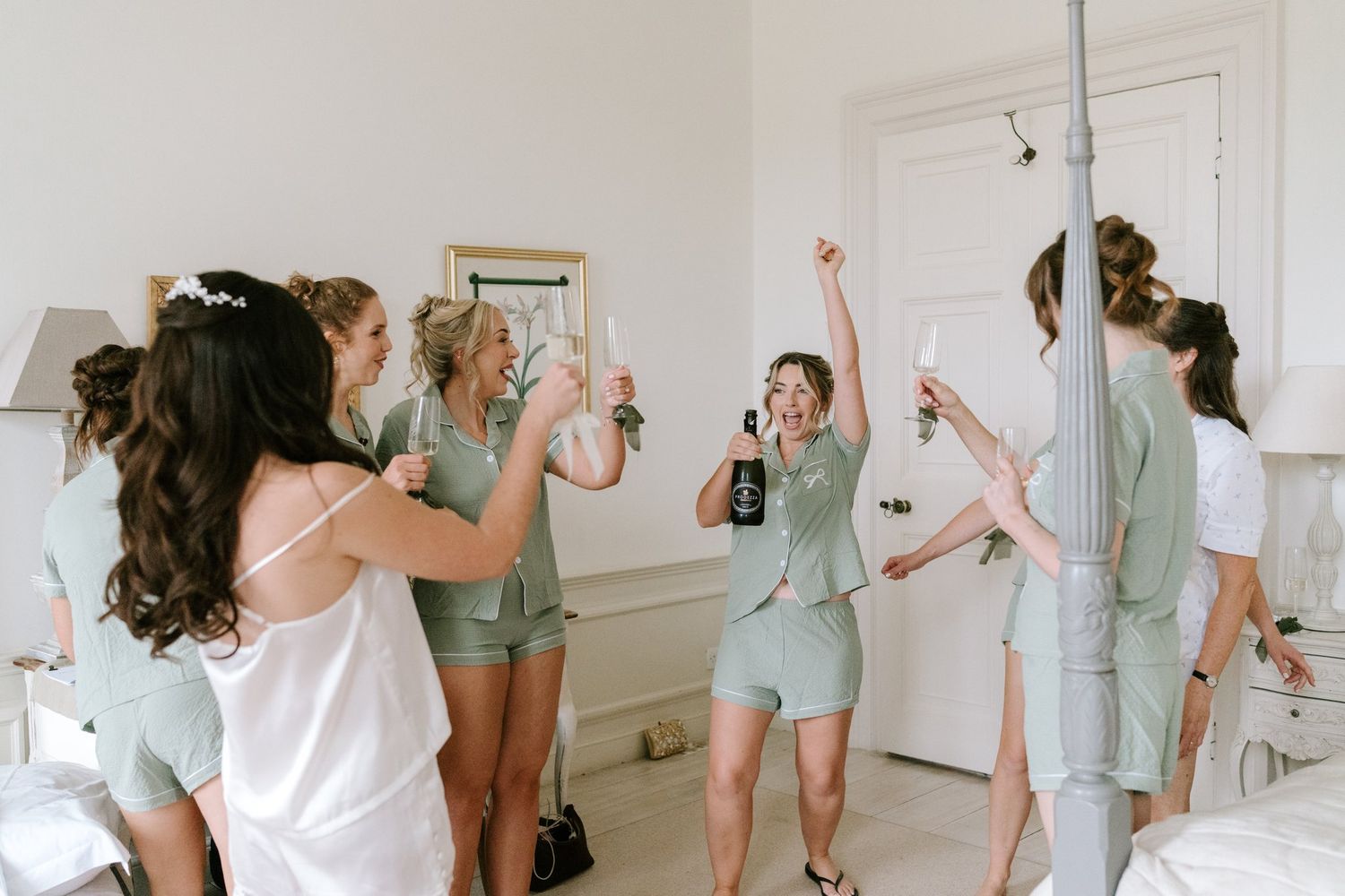 Bridesmaids in sage green pajamas celebrate with champagne in a bright white room before a wedding.