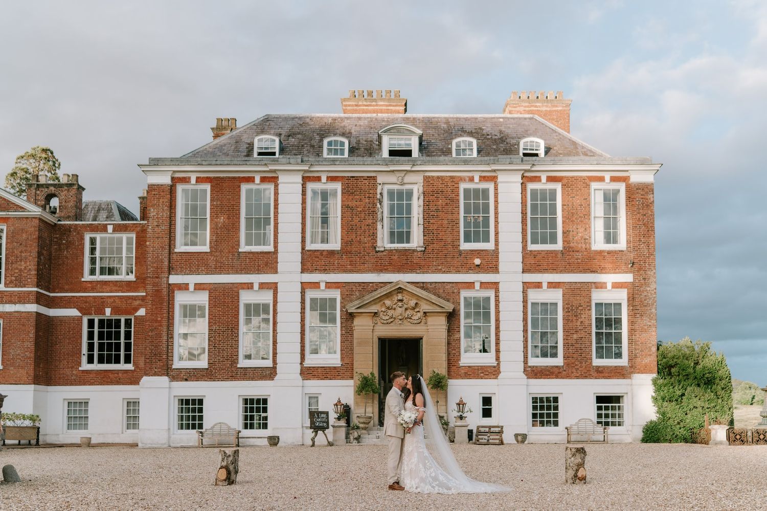 Bride and groom embracing outside a grand Georgian red brick manor house wedding venue on a cloudy day.