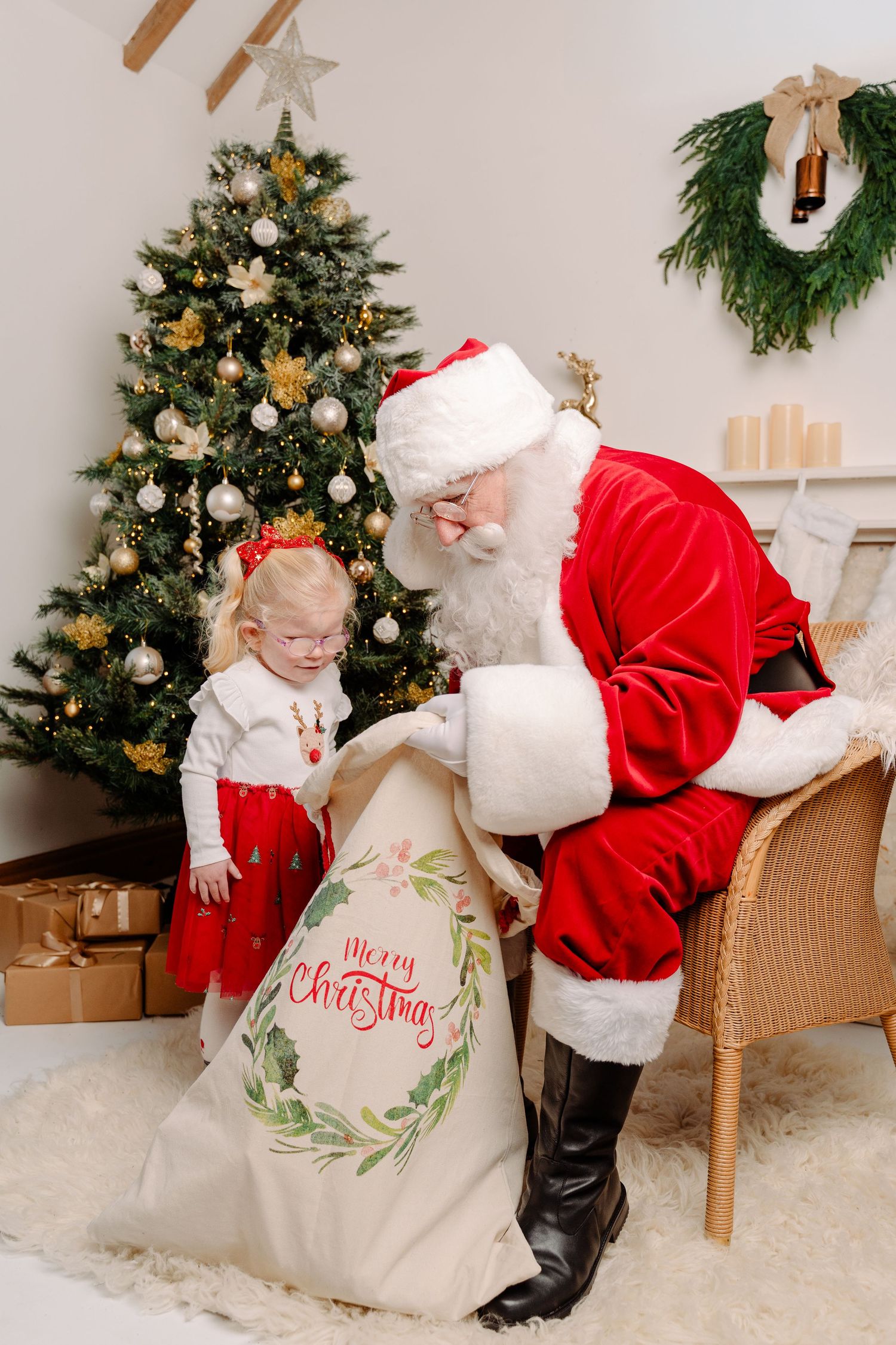 Santa Claus opens a Merry Christmas gift sack for a young girl in red dress beside a decorated Christmas tree.