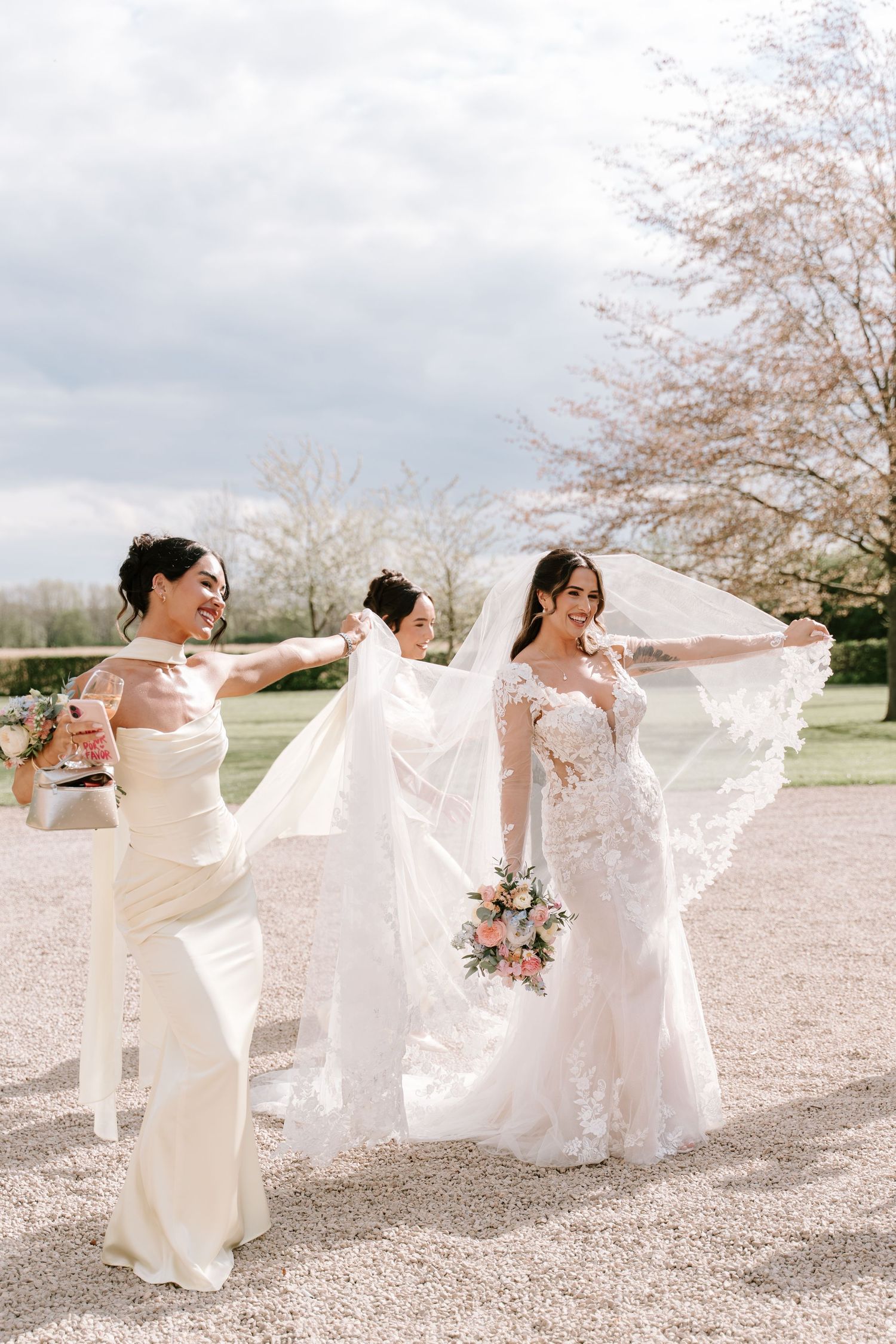 Bride in lace gown with flowing veil held by bridesmaids in white dresses, posing outdoors near cherry blossom trees.