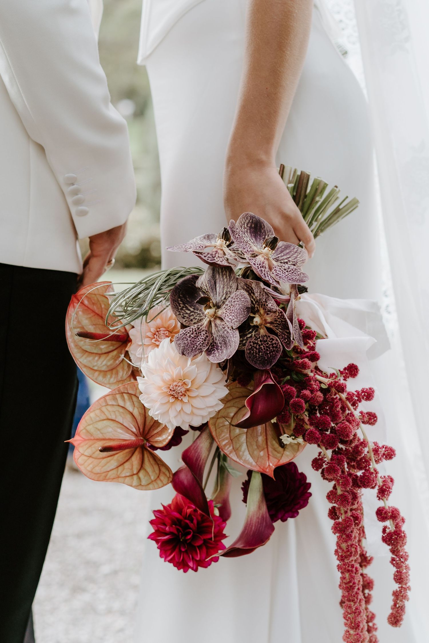 Bride holding a lush wedding bouquet with dahlias, anthuriums, orchids, and cascading amaranthus in deep burgundy and blush tones.