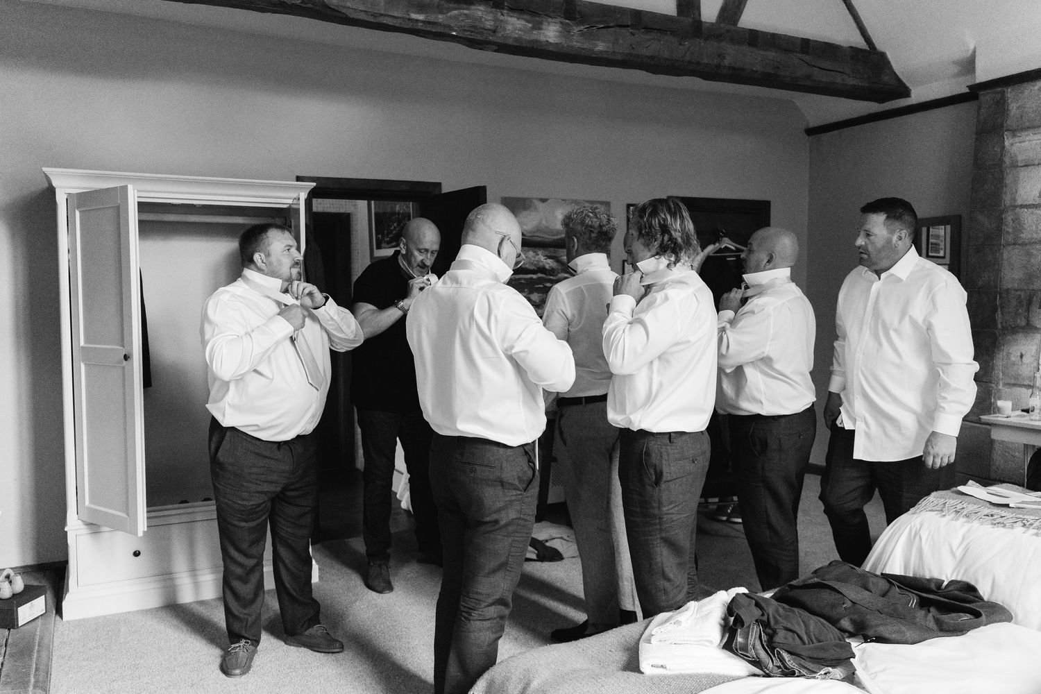Groomsmen helping each other with ties in a rustic bedroom while preparing for a wedding ceremony.