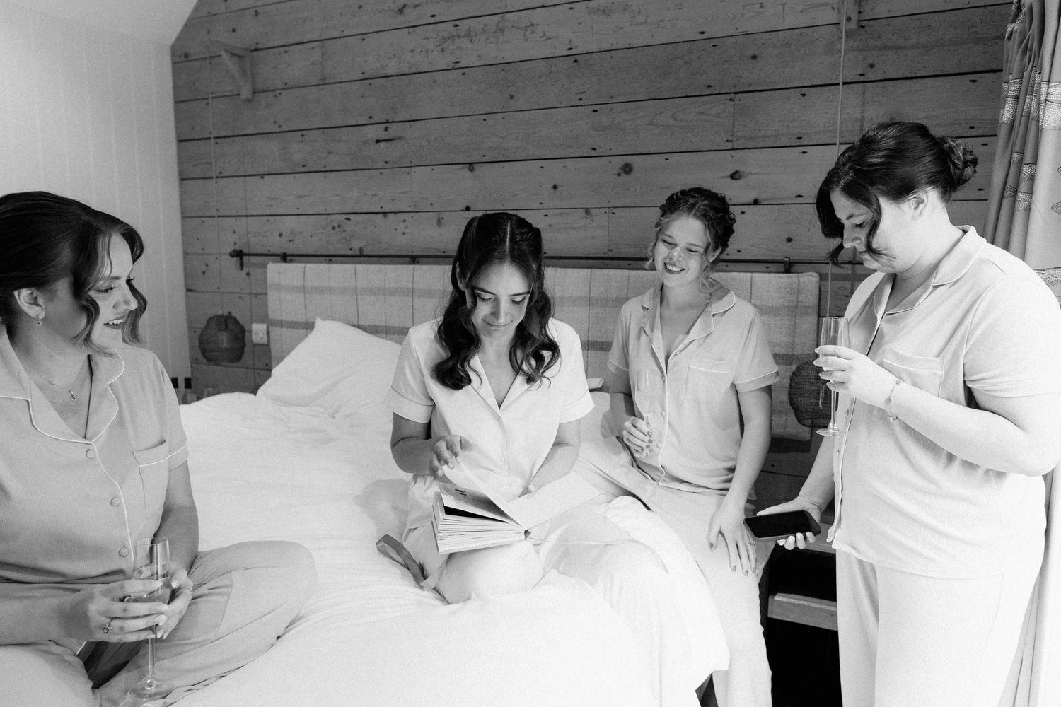 Bridesmaids in white pajamas gather on a bed, laughing and sharing moments together in a cozy rustic bedroom.